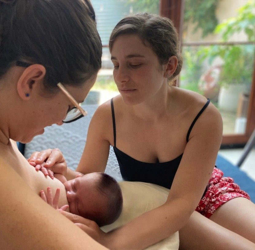 A woman is breastfeeding a newborn baby while another woman, her lactation consultant, looks on, sitting nearby in a cozy room with large windows and greenery outside.