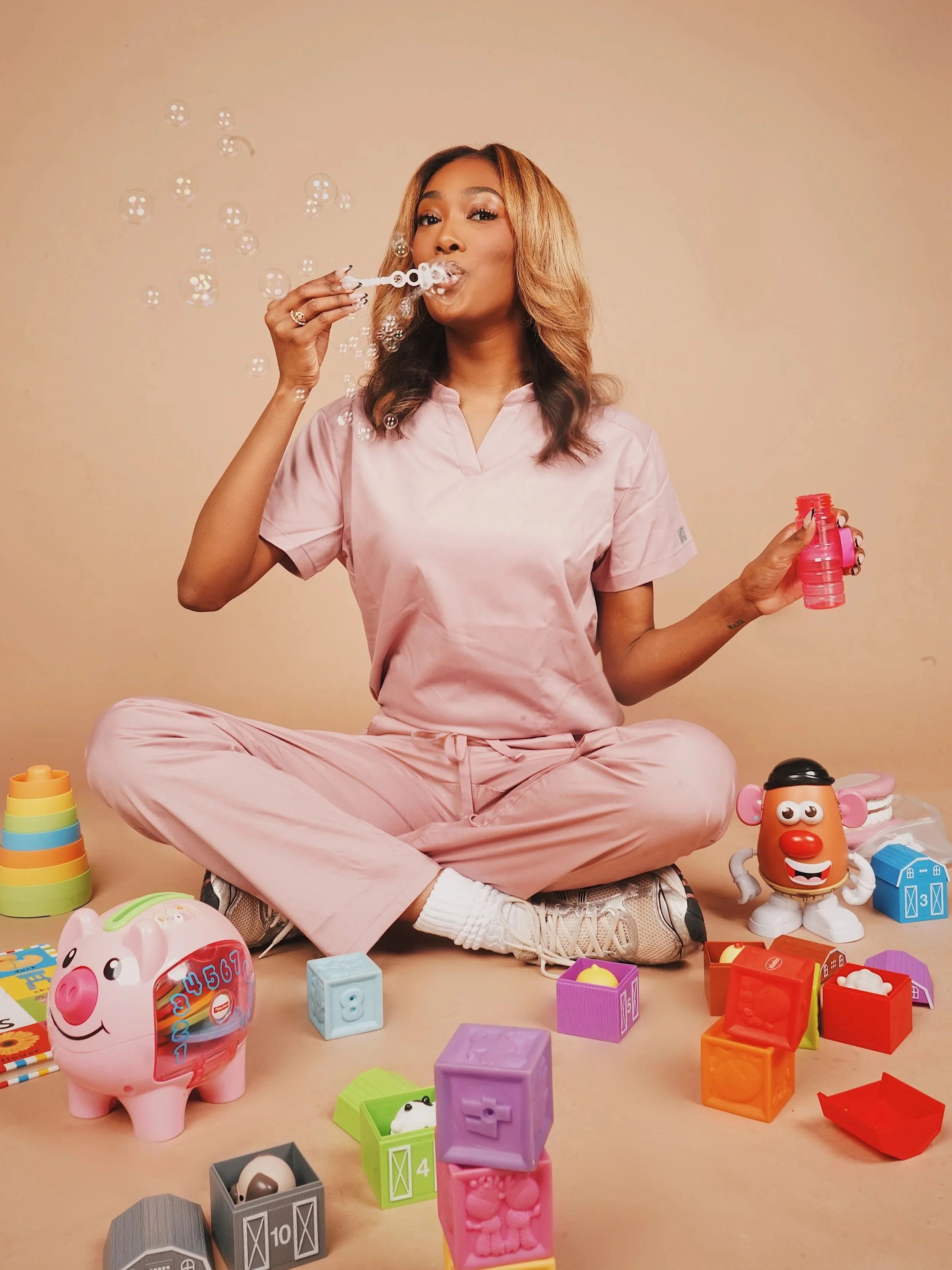 A woman in pink pajamas sitting cross-legged on the floor, blowing bubbles with a bubble wand, surrounded by colorful toys and blocks.