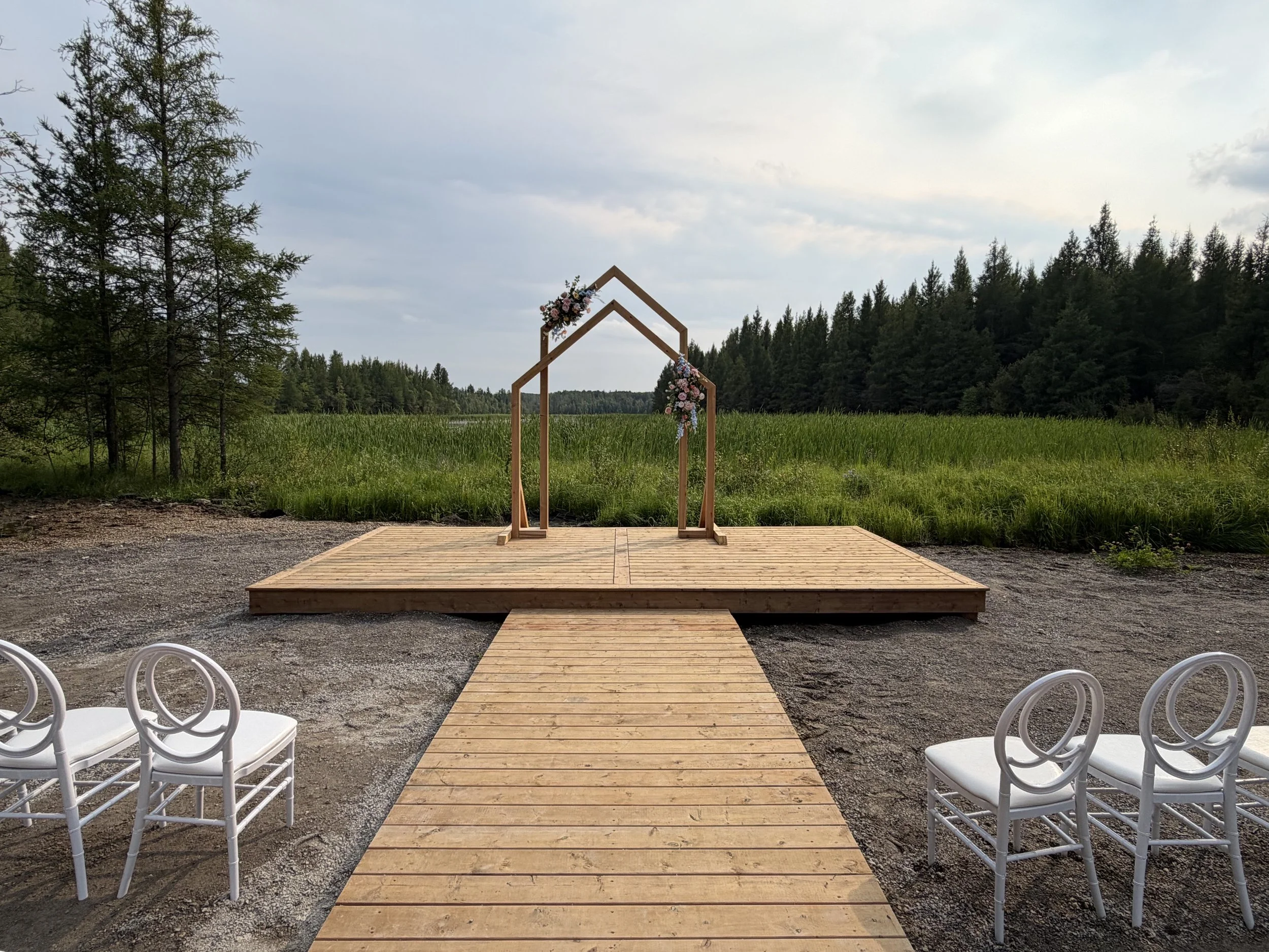 An outdoor wedding ceremony setup with a wooden aisle leading to a floral-decorated wooden arch, surrounded by green trees and a grassy field, under a cloudy sky.
