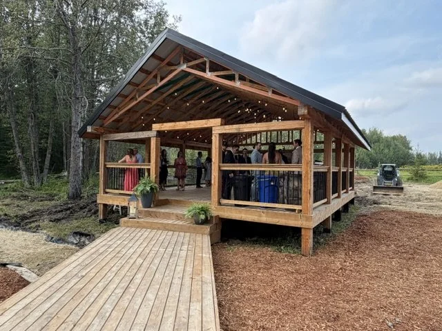 Wooden open-air pavilion with string lights, people socializing inside, a ramp leading up to it, and trees in the background