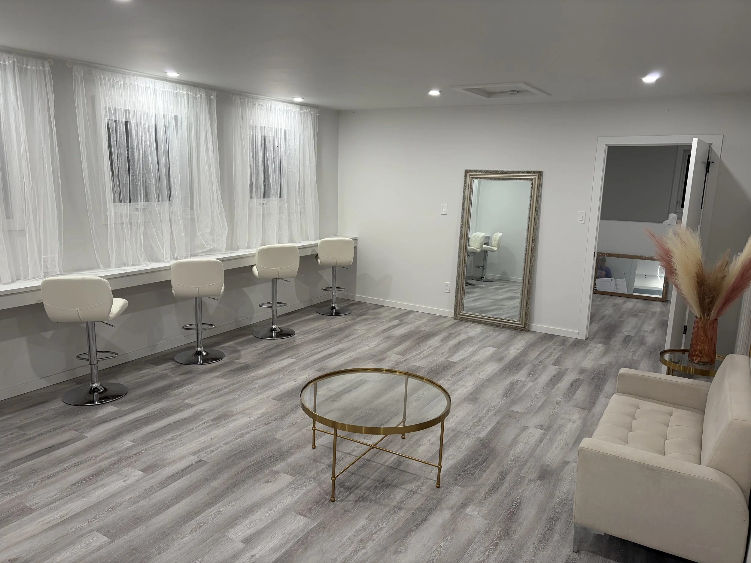 A modern, minimalist room with white walls, sheer curtains on large windows, and a row of white bar stools. Decorative items include a standing mirror with ornate frame, a round gold coffee table, and a cream-colored tufted sofa, with a vase of dried pampas grass in the corner.