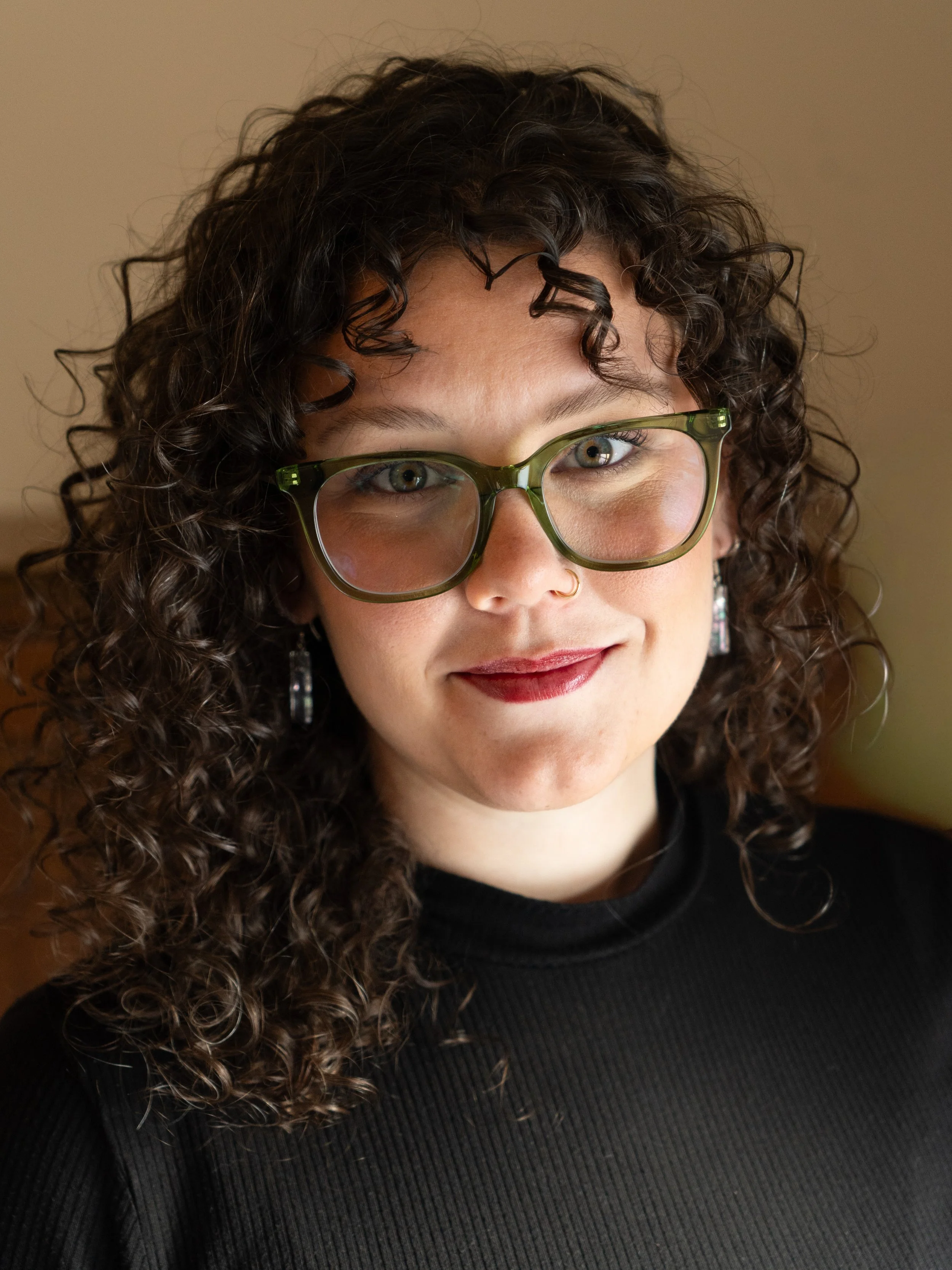 A woman with curly dark hair, wearing large green glasses, a nose ring, earrings, and a black top, smiling softly indoors.