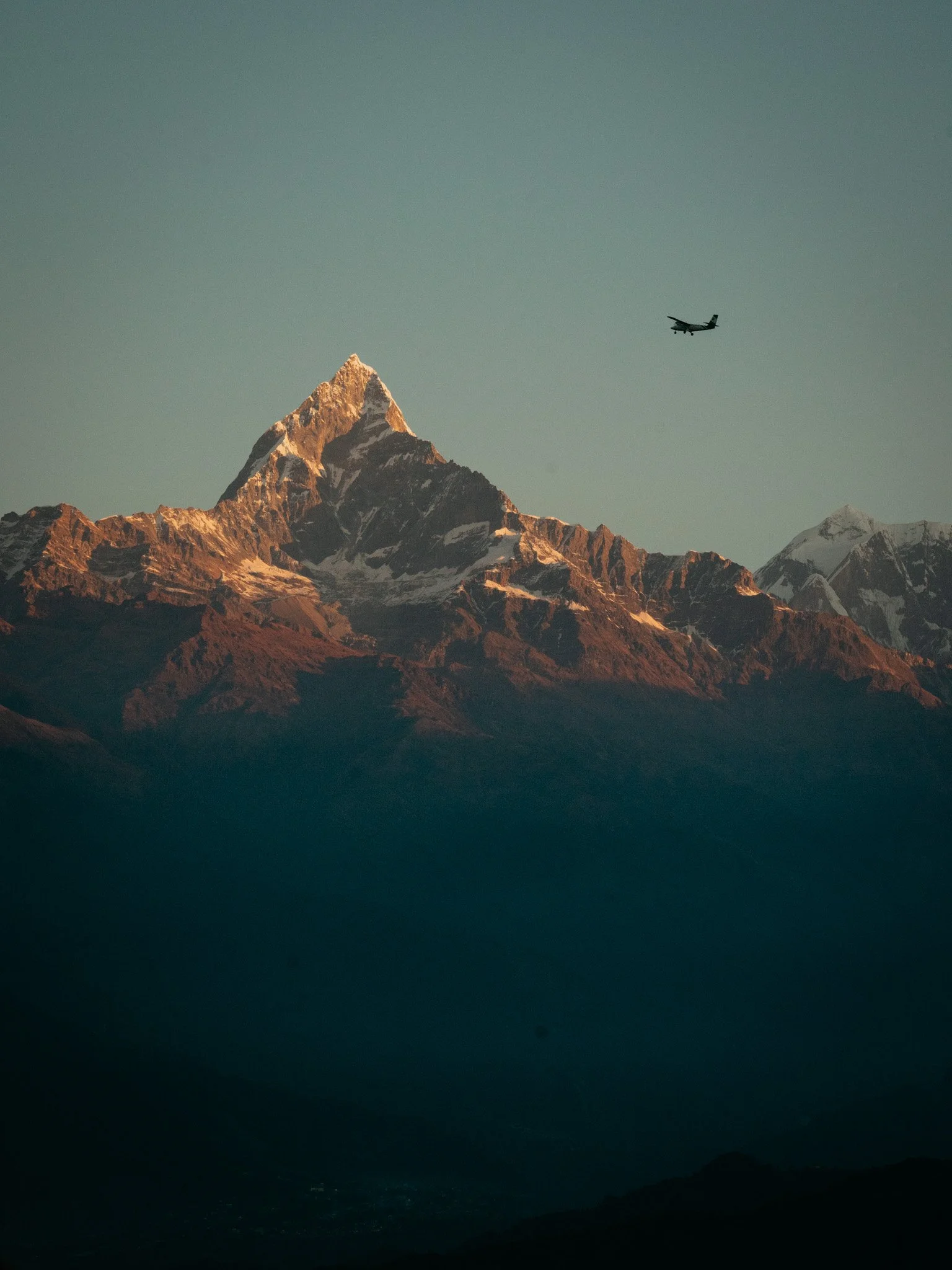 A snow-capped mountain peak with an airplane flying in the sky above it.