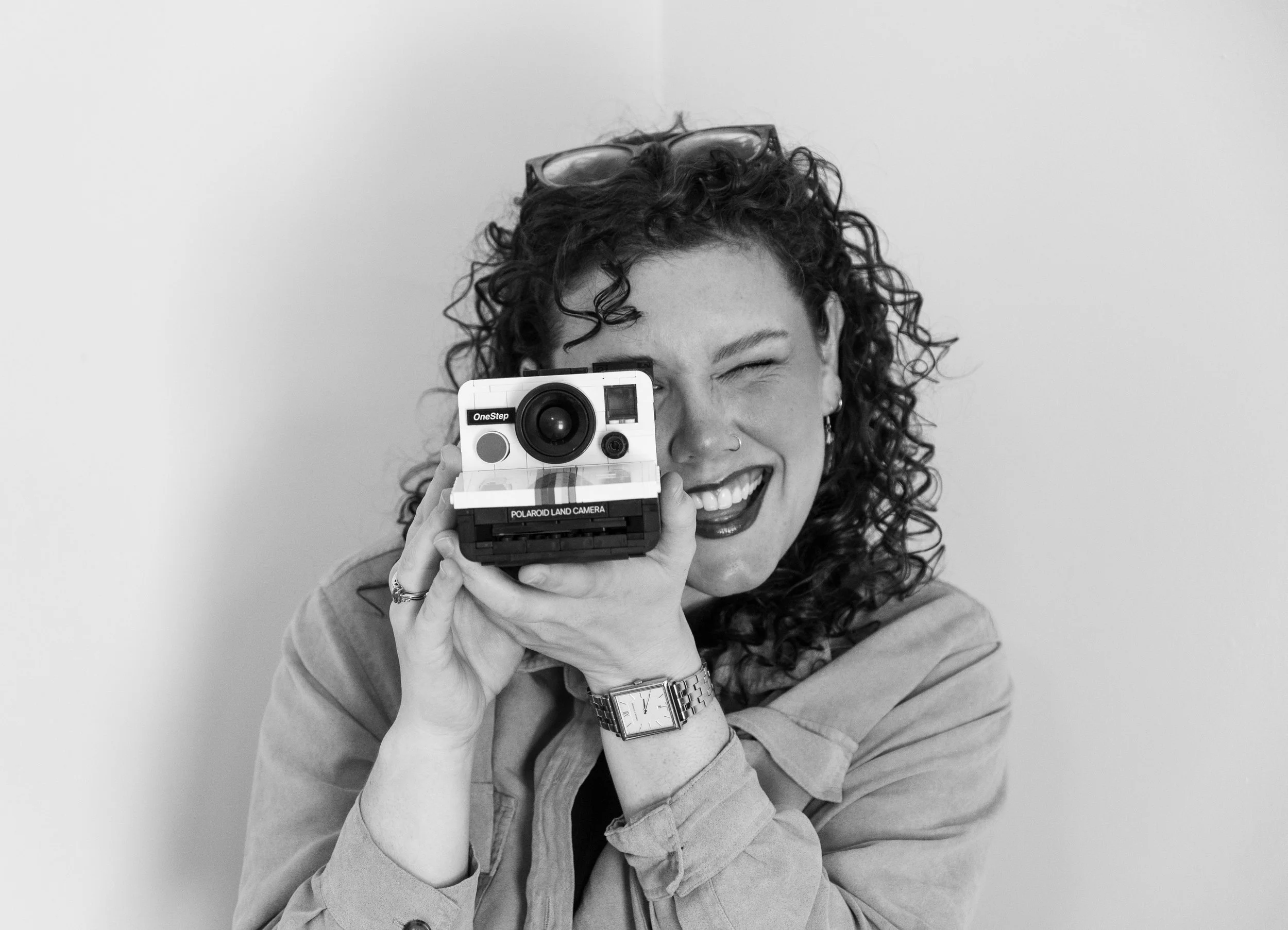 Black and white photo of a woman with curly hair smiling and holding a vintage Polaroid Land Camera up to her face.