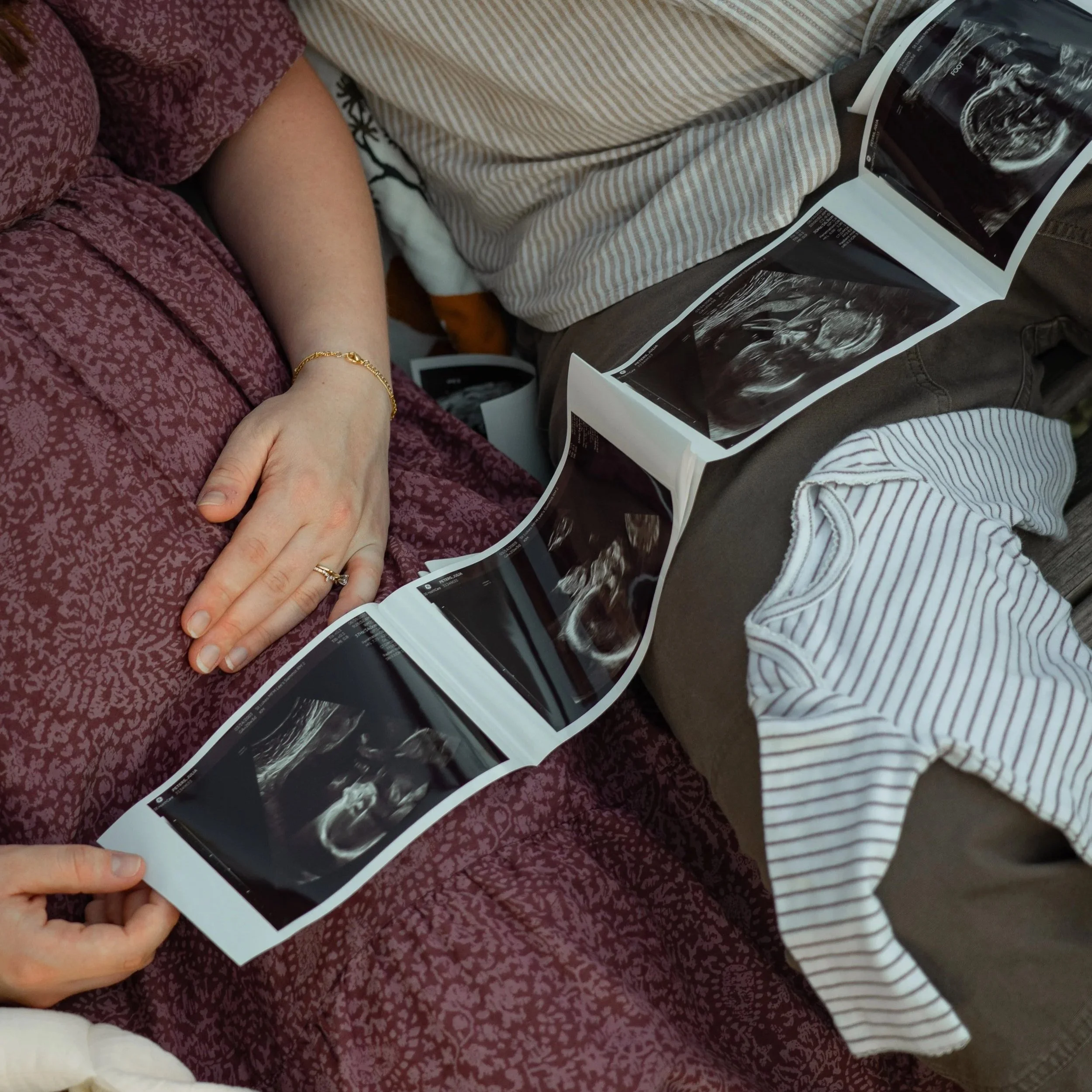 A couple sitting together, looking at ultrasound pictures of a fetus, with a woman wearing a burgundy patterned top and a man in a striped shirt, both holding the ultrasound sheet.