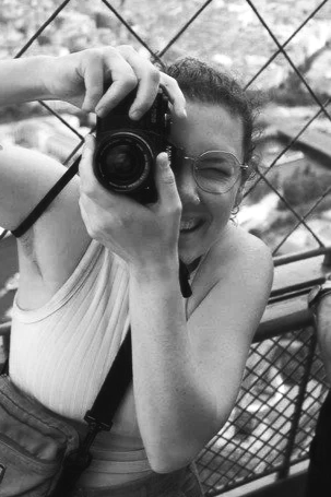 A woman taking a selfie with a camera. She is smiling, wearing glasses, and appears to be outdoors near a metal fence.