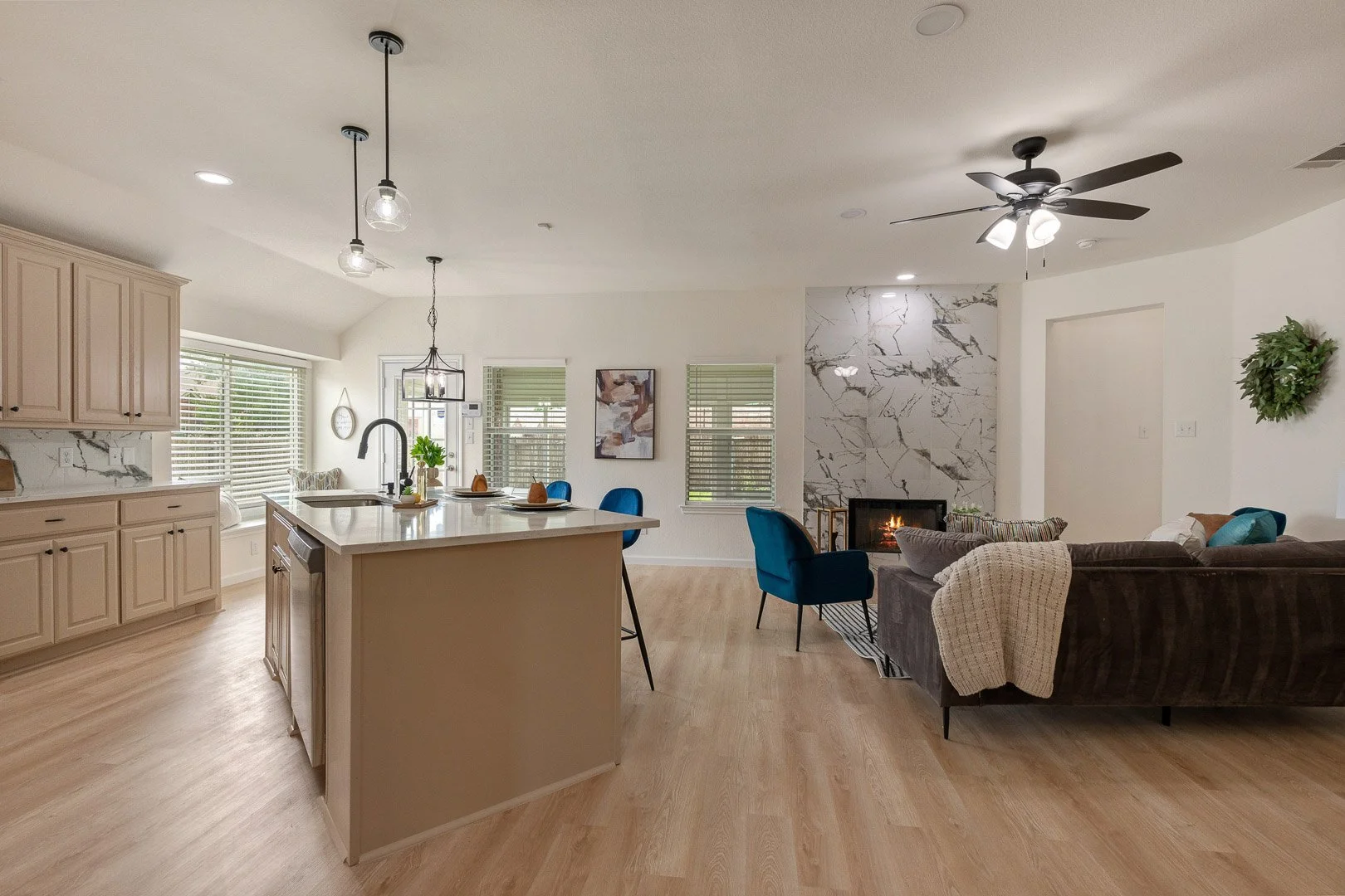 Open-concept living room and kitchen area with light hardwood floors, a gray sectional sofa, a modern fireplace with marble surround, a kitchen island with barstools, and large windows.