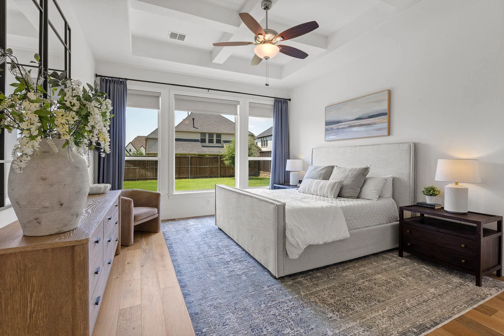Bright bedroom with large windows, white walls, beige upholstered bed, dark wooden nightstands with lamps, a colorful painting above the bed, a dresser with a large vase of flowers, a beige armchair, a ceiling fan, and wood flooring with a patterned 