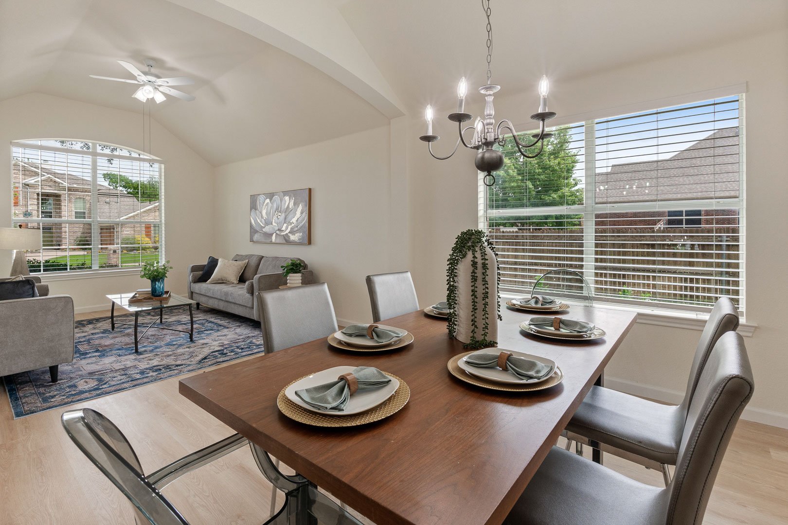 Dining area with a wooden table set with plates, napkins, and a vase with greenery, adjacent to a living room with sofas, a coffee table, and a large window with blinds, in a modern home.
