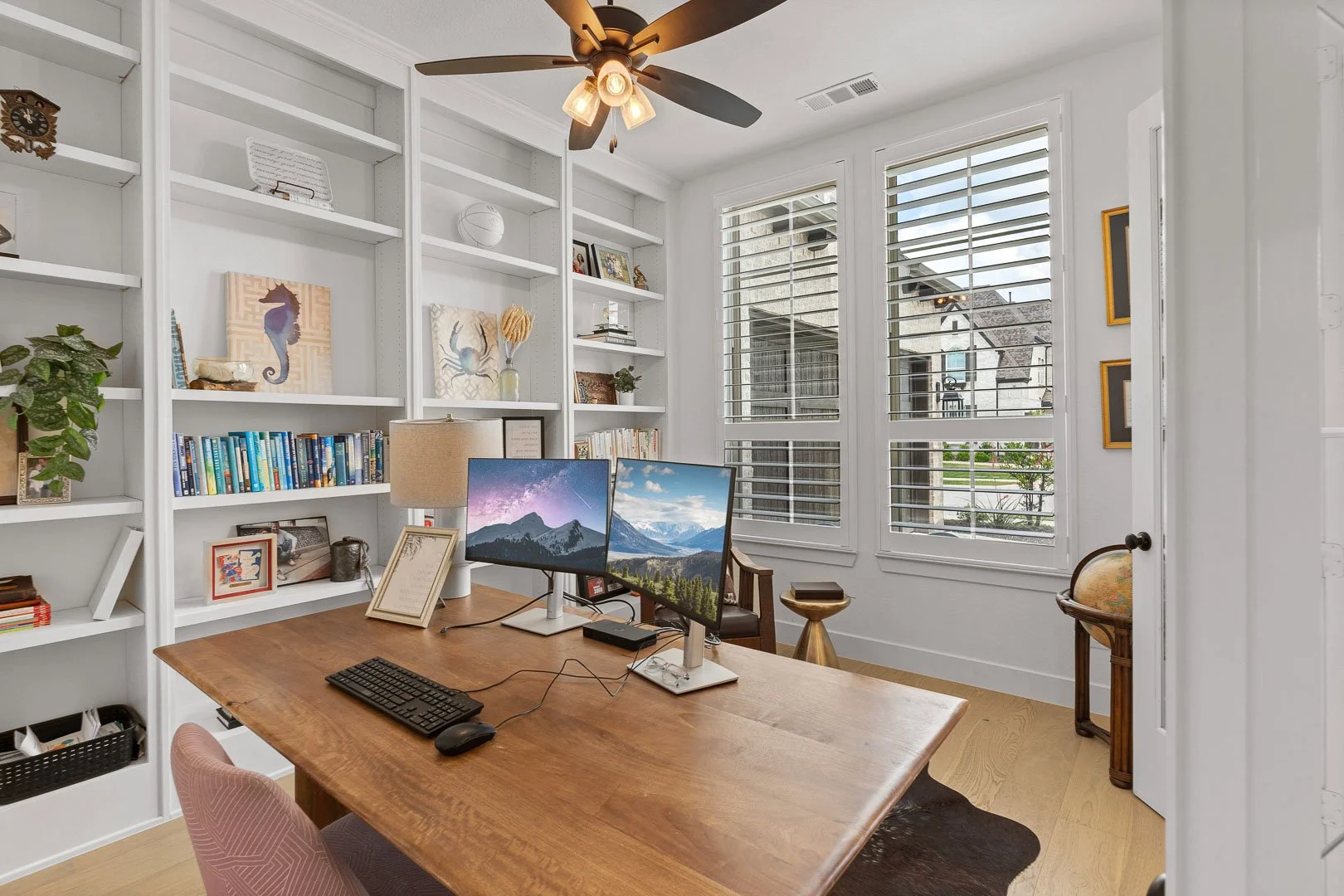 A home office with a large wooden desk, dual computer monitors, a pink chair, a lamp, and pictures on the shelves. Windows with white blinds let in natural light. The room has a ceiling fan and white walls.