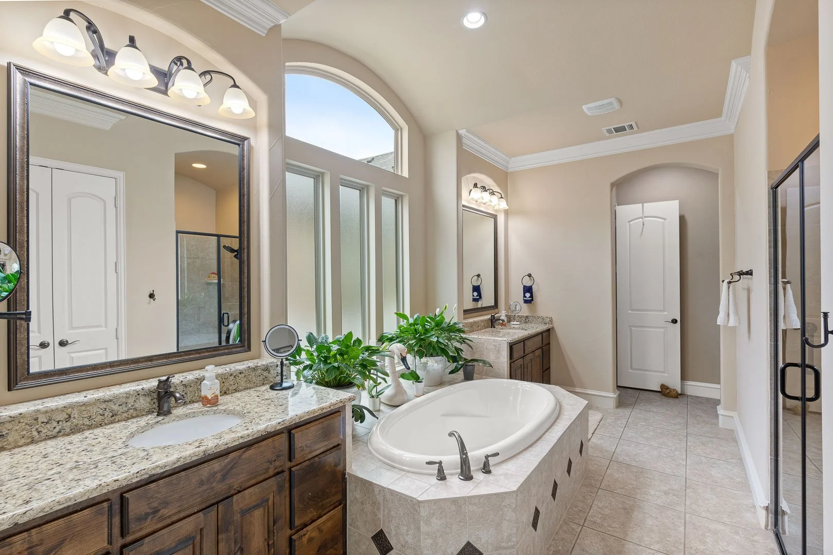 Bathroom with double vanity, large mirror, corner bathtub with plants, shower with glass doors, and off-white walls.