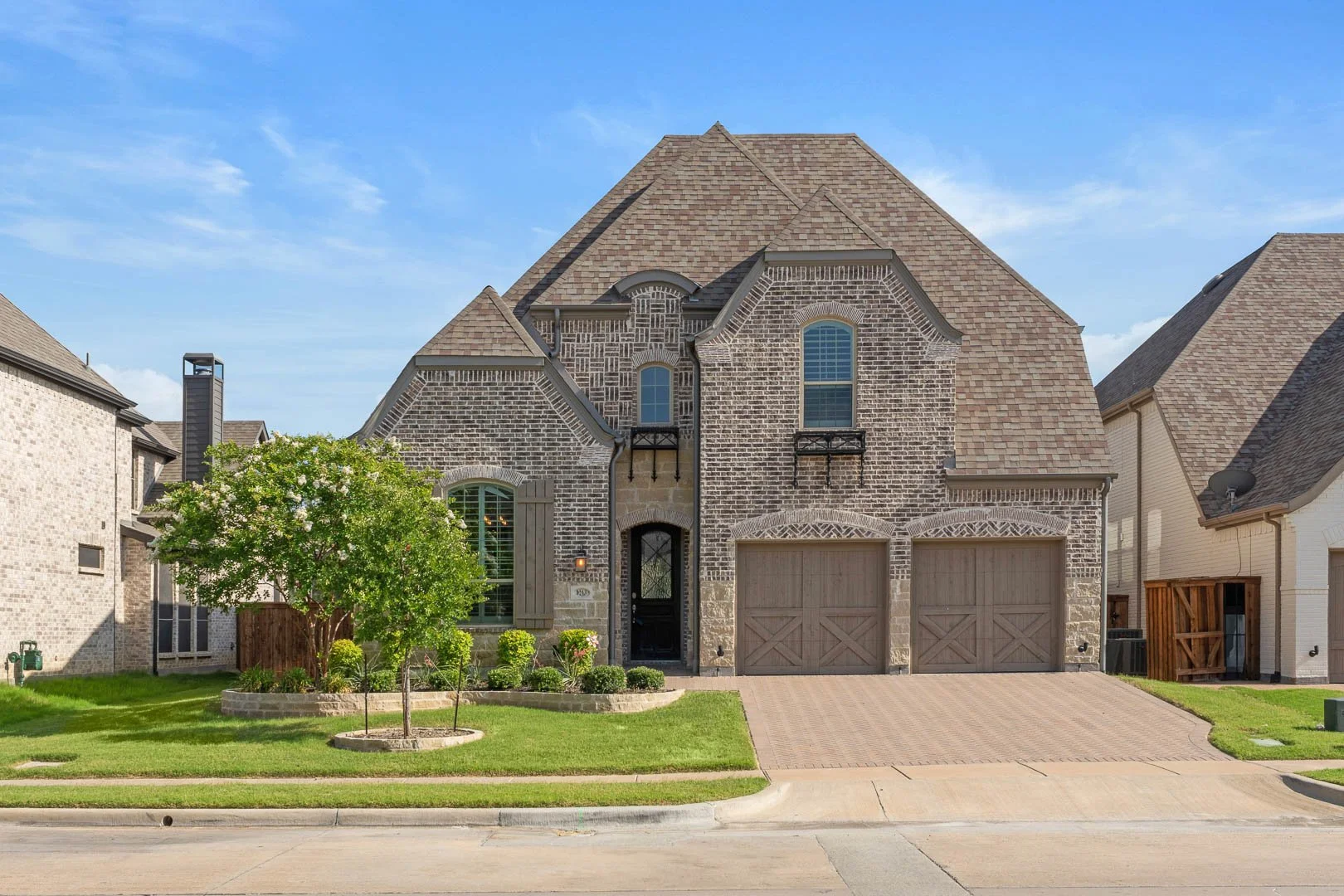 A two-story brick house with a front lawn, driveway, two garage doors, and a small landscaped garden with a tree and shrubs, under a blue sky.