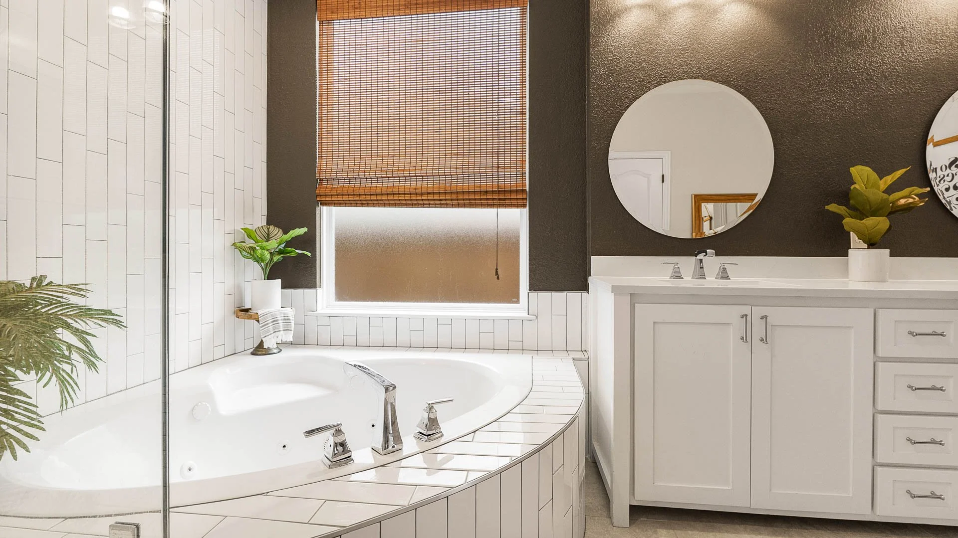 Bathroom with a white bathtub next to a frosted window with a bamboo shade, white cabinets, and two round mirrors on a dark gray wall, decorated with green plants.