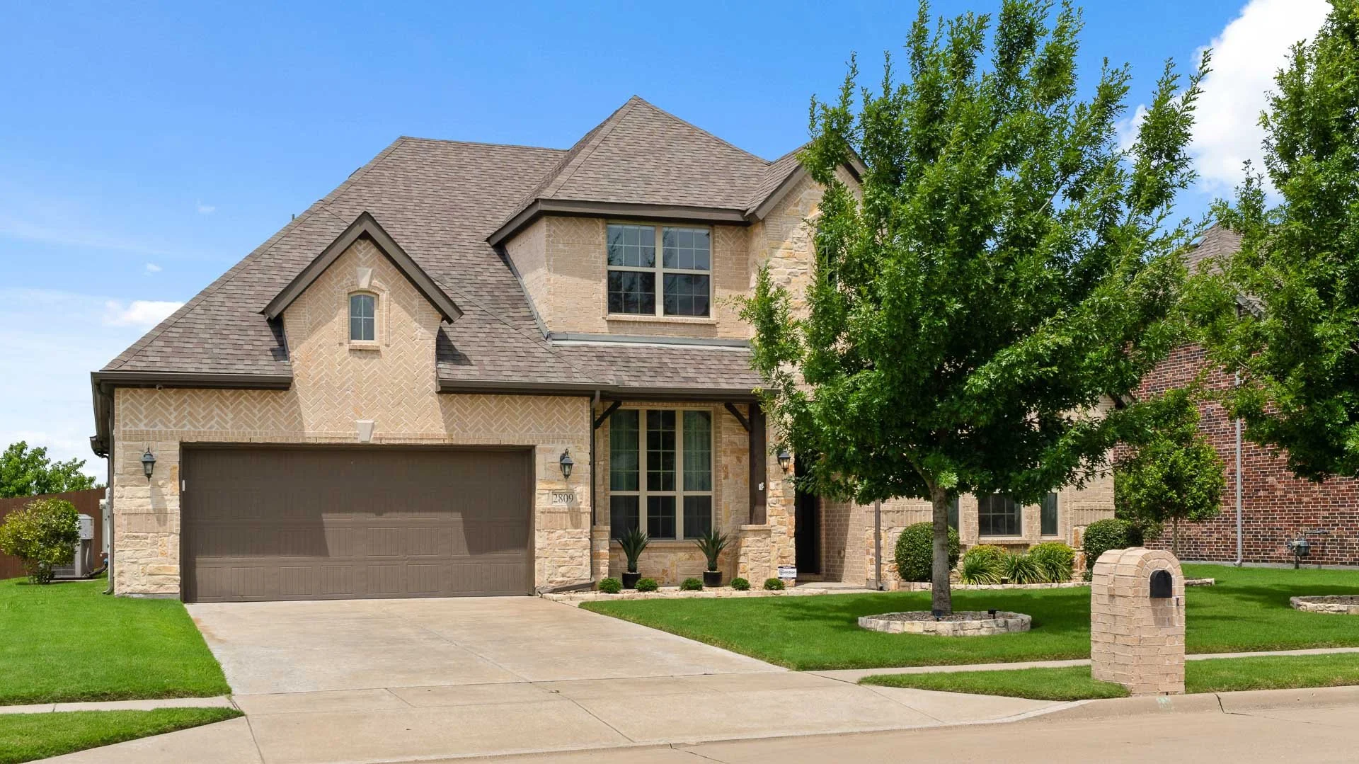 A two-story suburban house with beige brick and stone exterior, brown roof, green lawn, and a tree in front. There is a driveway leading to a two-car garage and a mailbox in the yard.