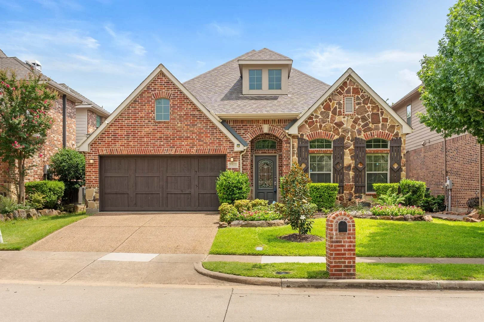Front view of a house with brick and stone exterior, a two-car garage, a black front door, and a well-maintained lawn with bushes and flowers, under a blue sky.