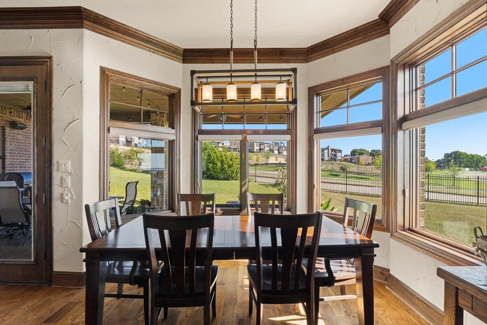 Dining room with large windows, wooden table, six chairs, and a modern chandelier, overlooking a green lawn with buildings in the distance.