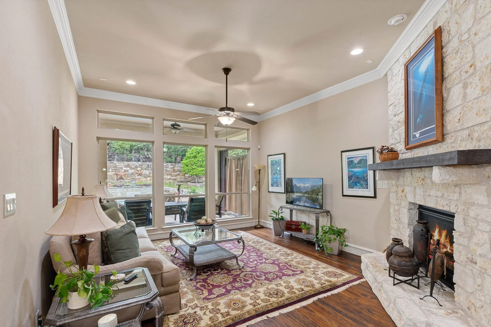 Living room with beige walls, fireplace with stone surround, framed artwork, ceiling fan, large windows showing outdoor patio with seating and garden, sofa, coffee table, plants, TV, and rugs.