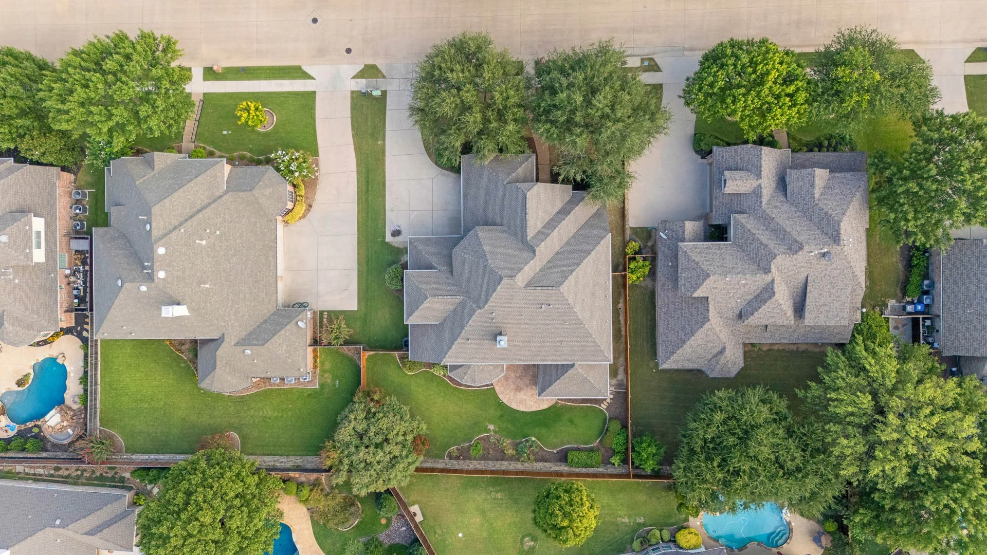 Aerial view of three neighboring houses with backyards, trees, lawns, driveways, and swimming pools in a suburban neighborhood.