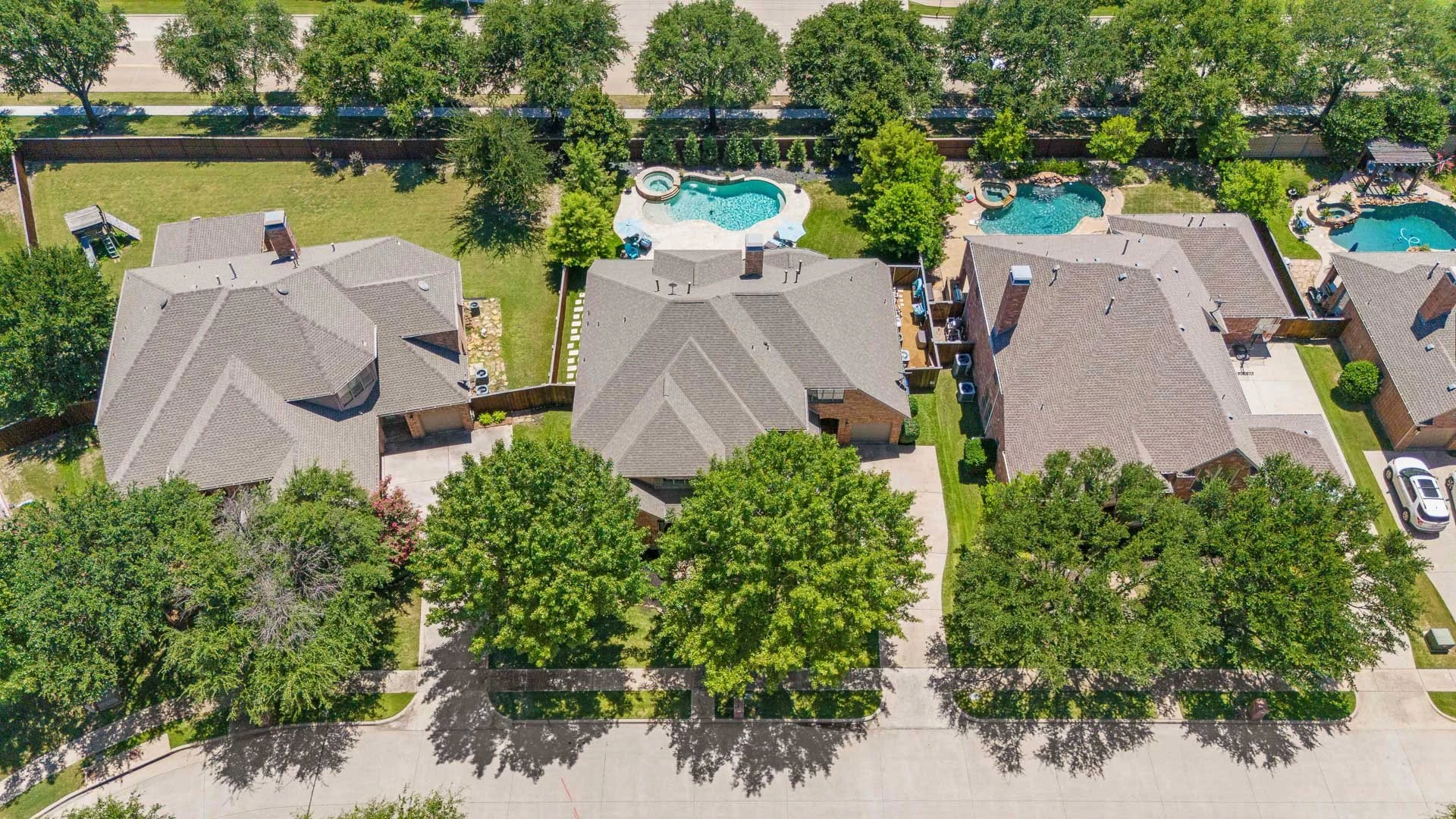 An aerial view of a residential neighborhood showing three houses with gray roofs, backyards with trees, and swimming pools. The houses are separated by fences, and there is a street at the bottom of the image.