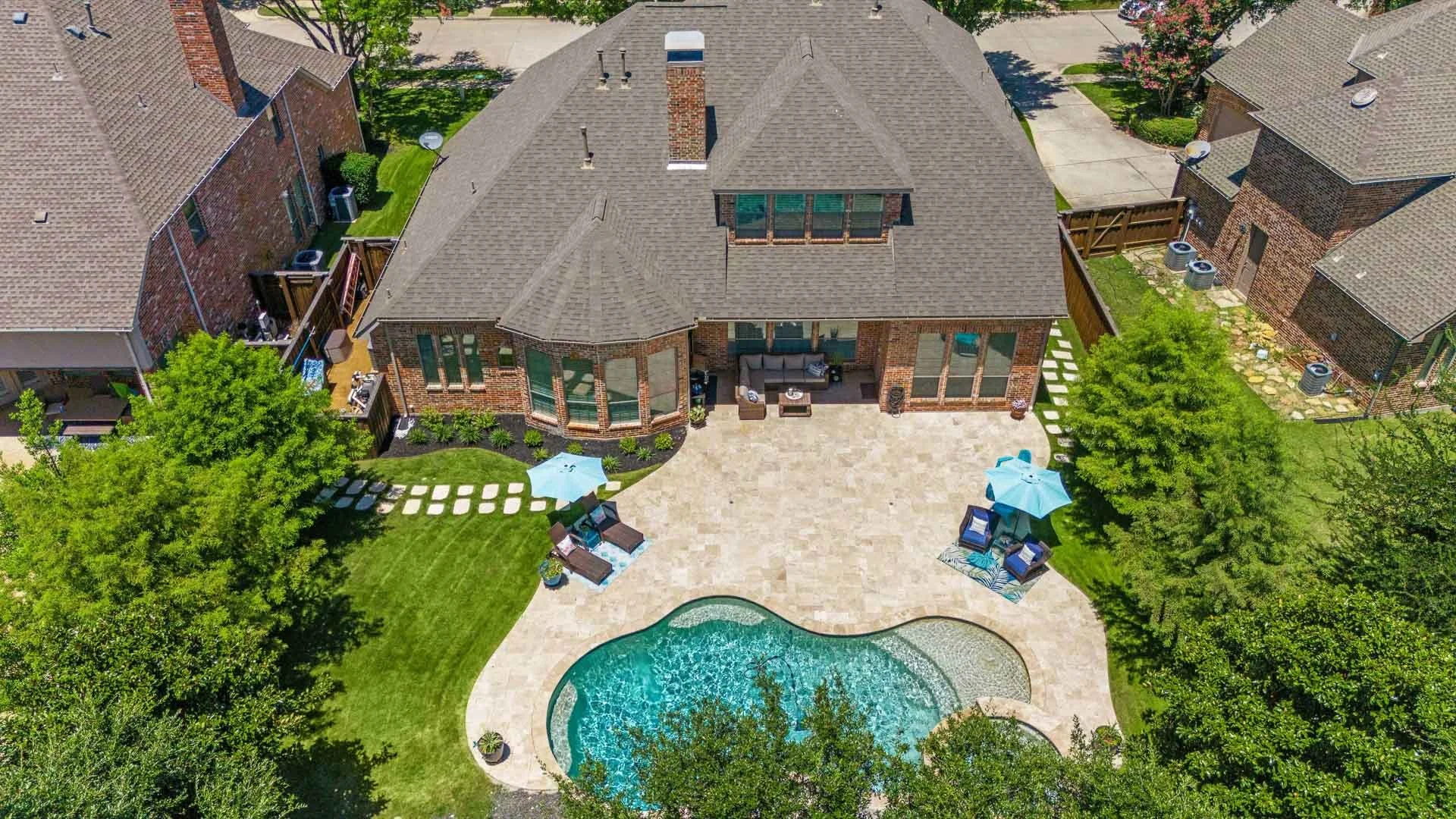 Aerial view of a brick house with a backyard featuring a kidney-shaped swimming pool, outdoor seating areas with umbrellas, lush green grass, and surrounding trees.