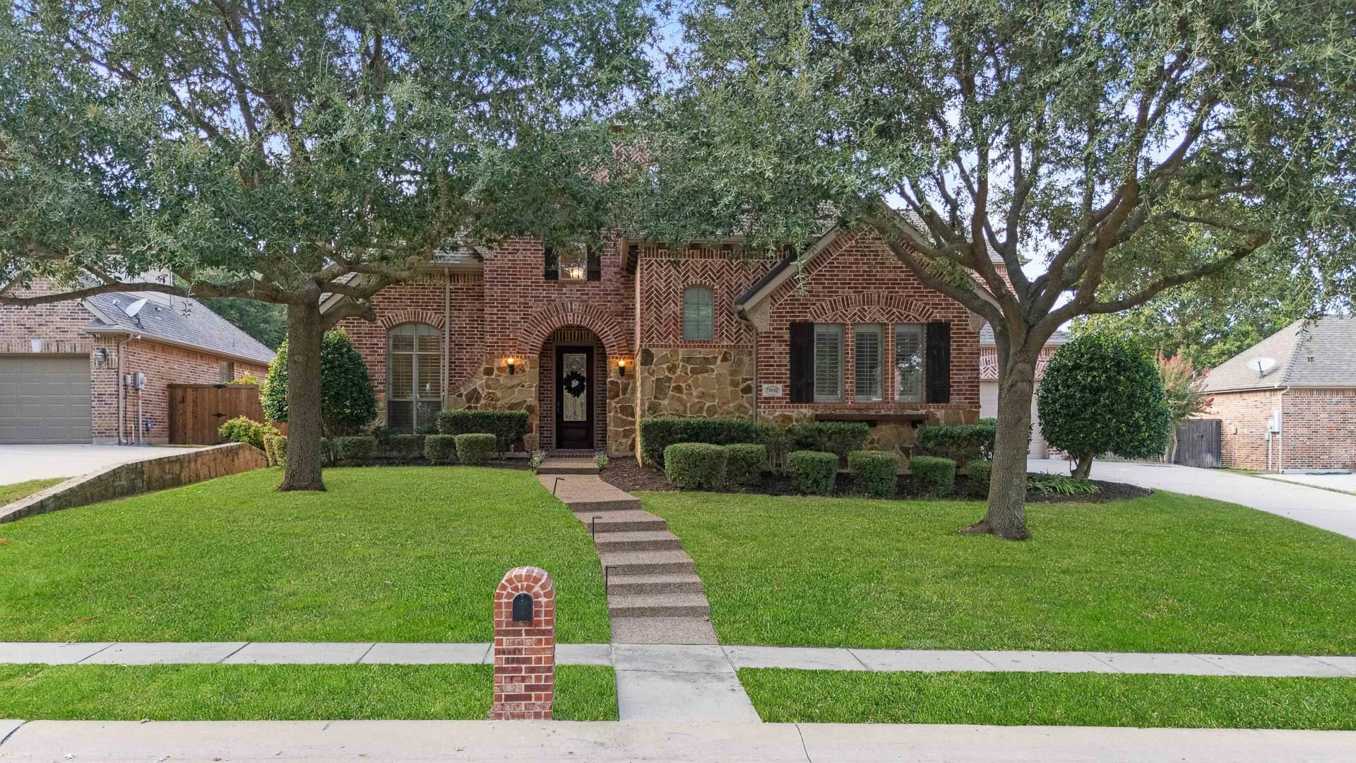 Front view of a brick house with stone accents, two large trees, a lawn, and a sidewalk with a brick mailbox.