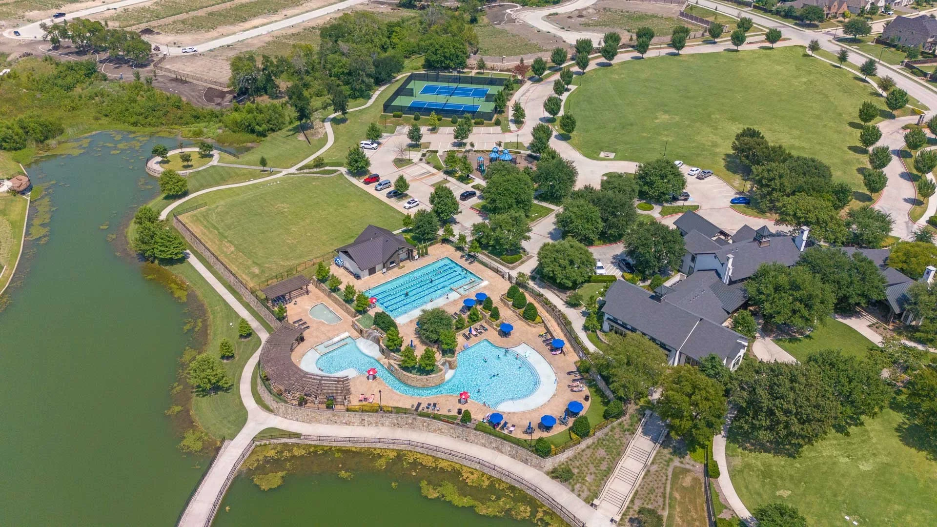 Aerial view of a community recreational area with a swimming pool, hot tub, tennis courts, and a pond, surrounded by walking paths, benches, and trees.