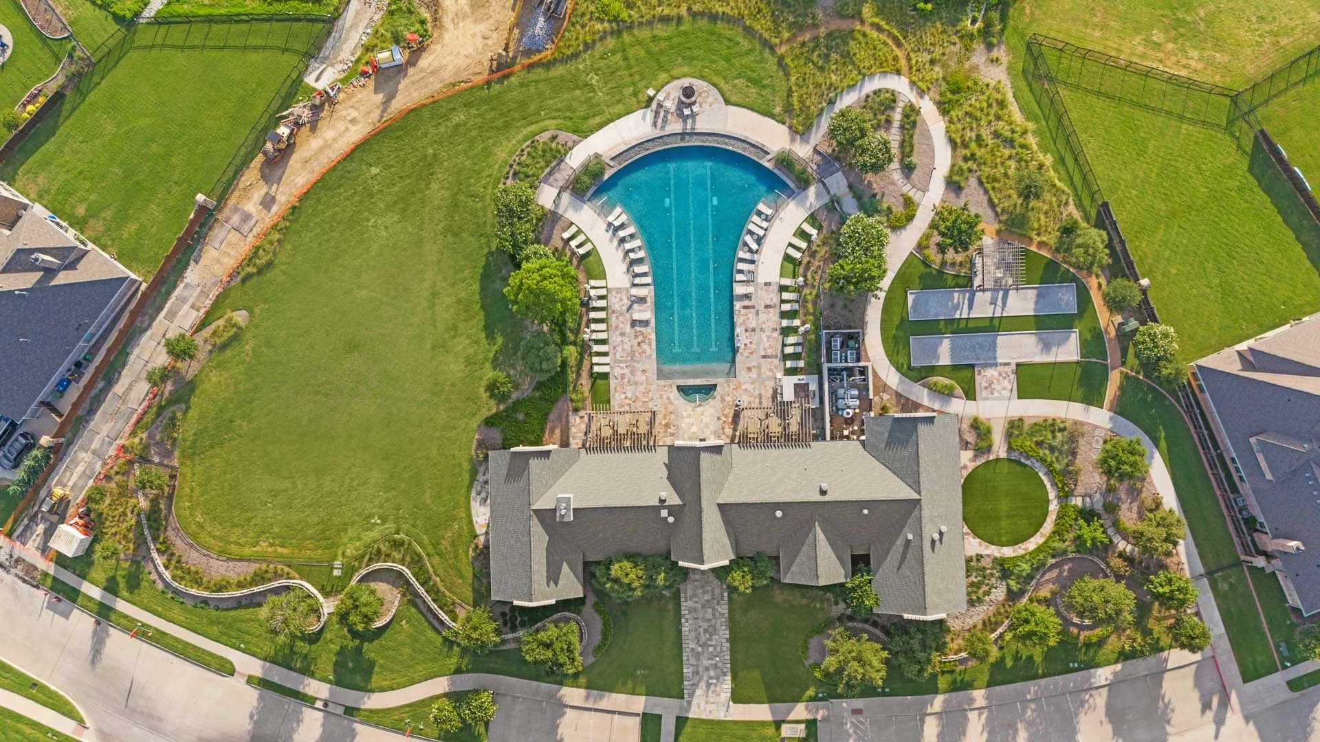 Aerial view of a backyard with a large swimming pool, surrounded by lounge chairs and greenery, with pathways and a house at the bottom.