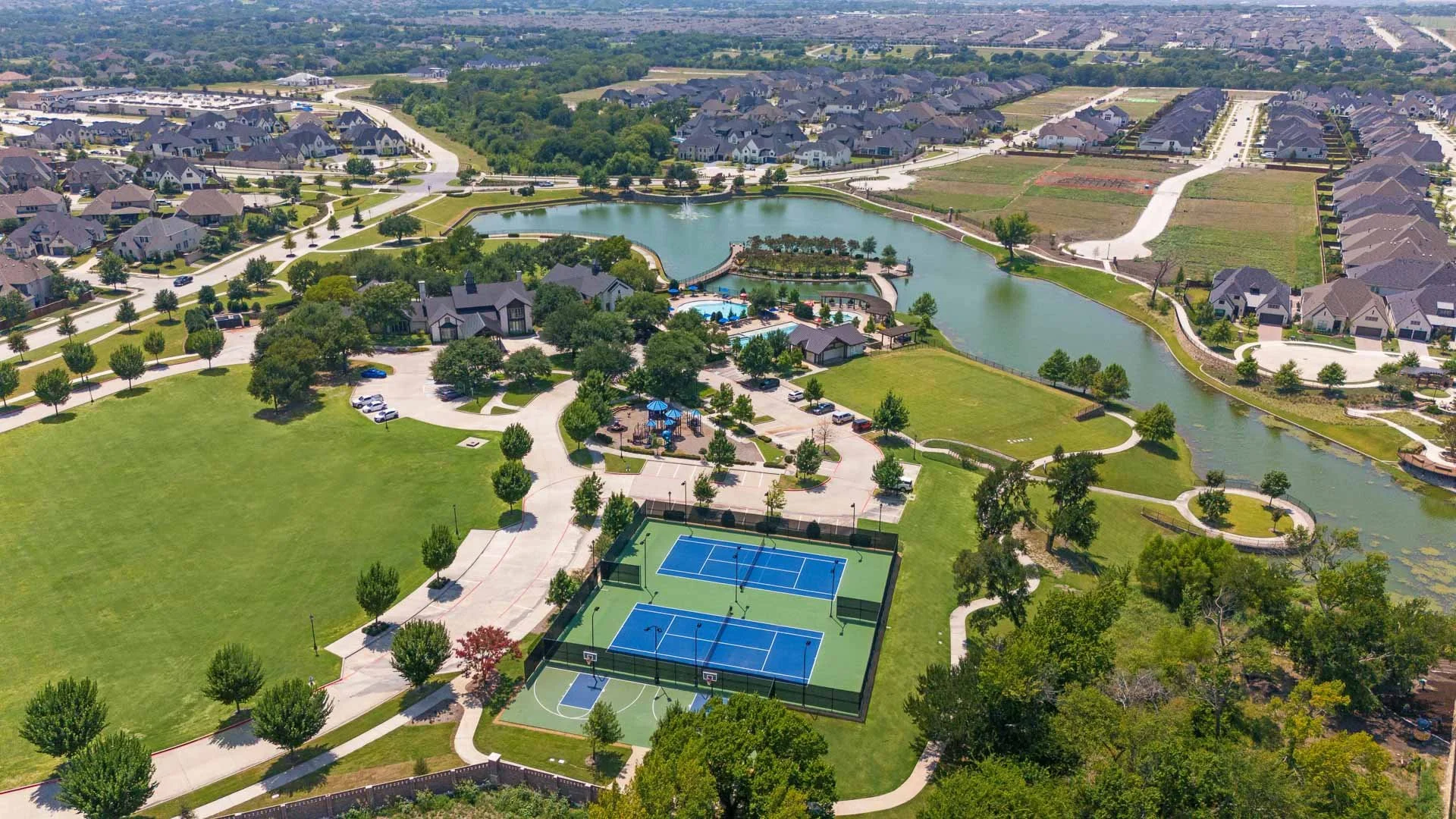 Aerial view of a residential community featuring a large pond, a playground with blue shade covering, multiple swimming pools, tennis courts, and a wide grassy field with trees.