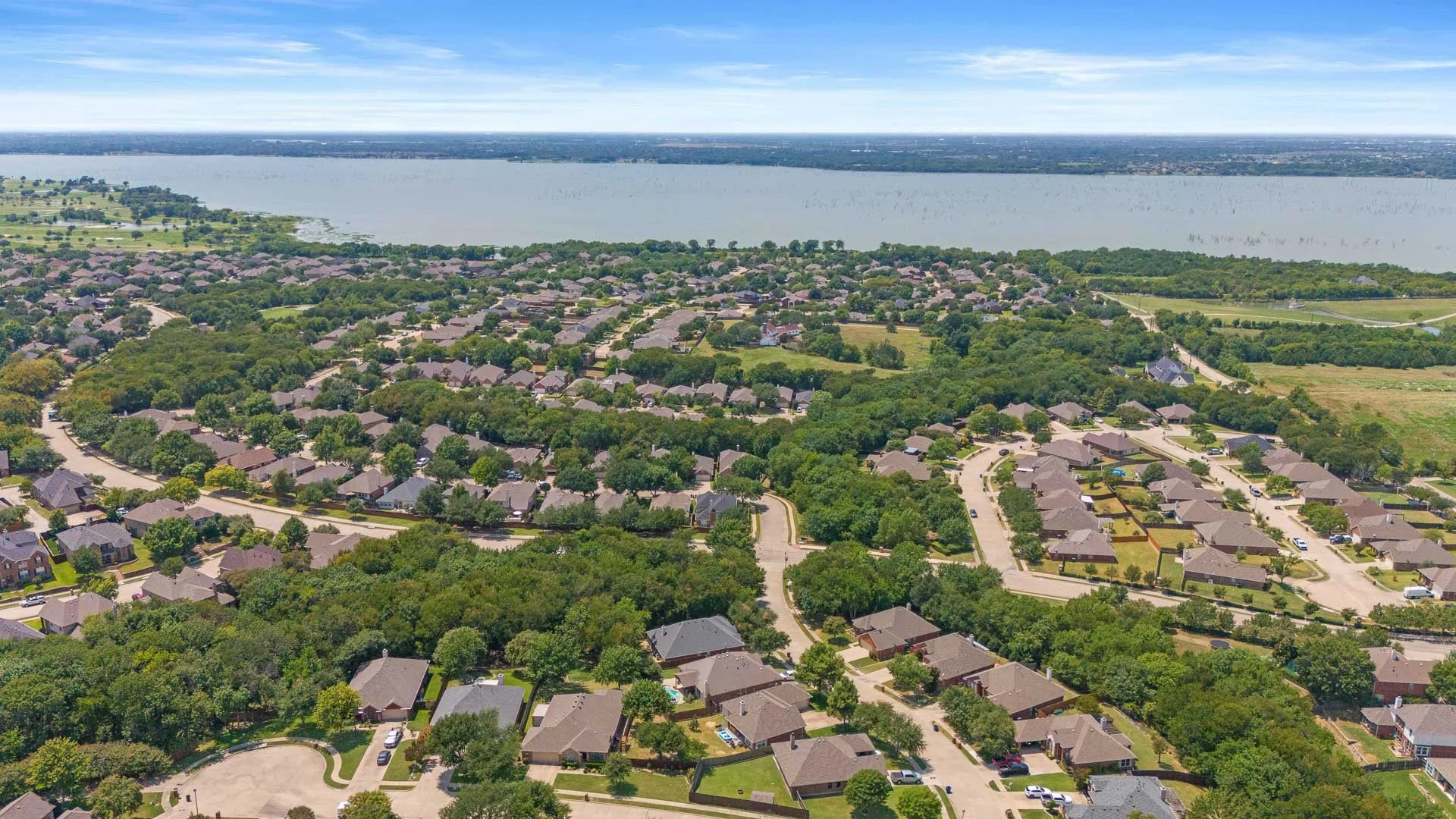 Aerial view of a suburban neighborhood near a large body of water, with houses, trees, and winding roads.