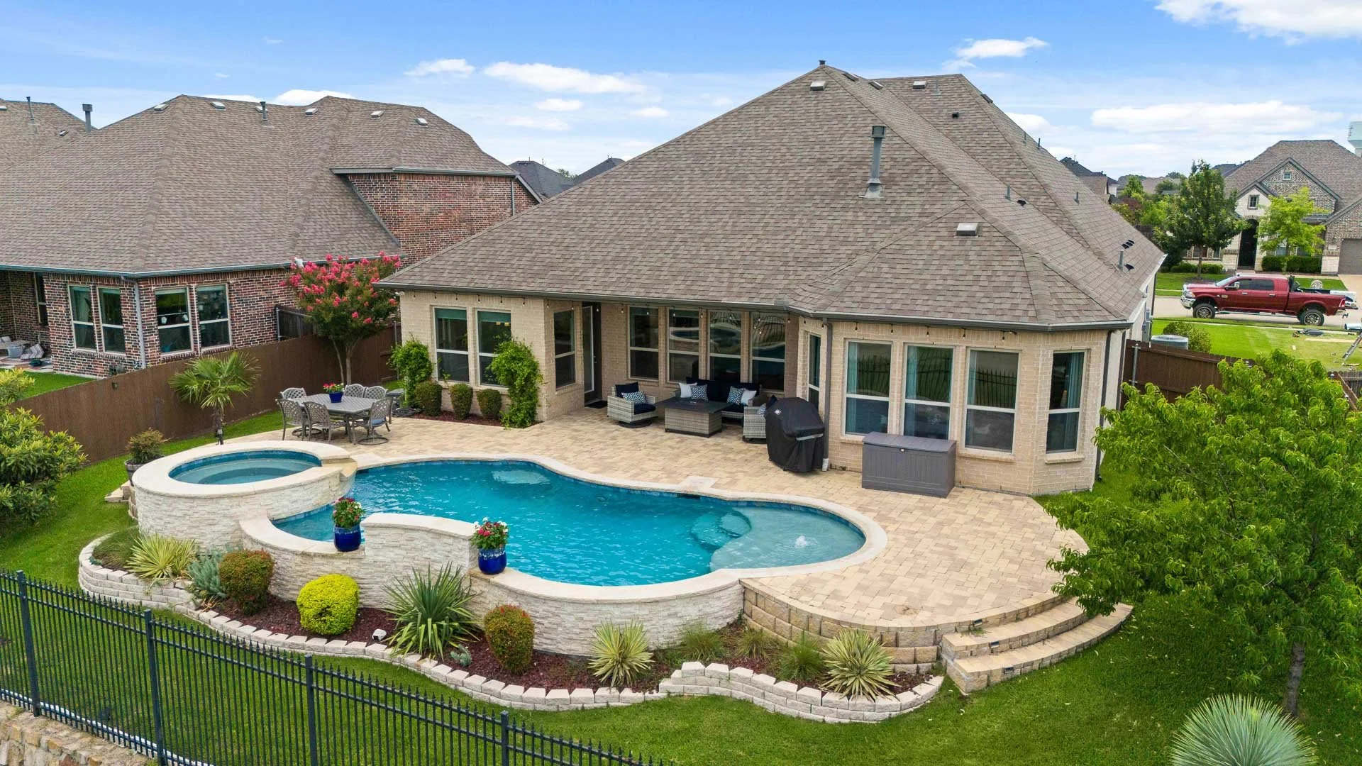 Backyard of a house with a swimming pool, hot tub, patio furniture, and landscaped garden. Large house with multiple windows and a sloped roof. Recent neighborhood with other houses and a street with parked truck visible.