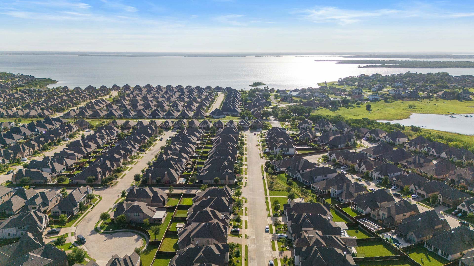 Aerial view of a suburban neighborhood overlooking a large body of water with houses, green lawns, and streets.