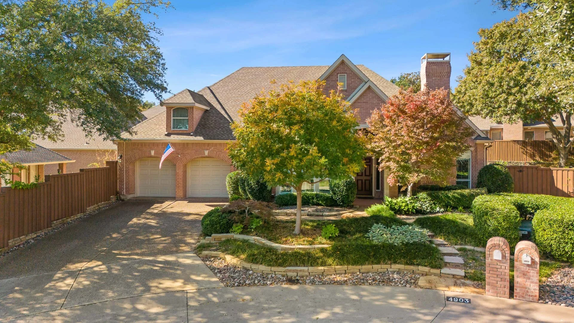 Front view of a two-story brick house with a driveway, two garage doors, and lush landscaping including trees, shrubs, and a flower bed. An American flag is visible near the garage.