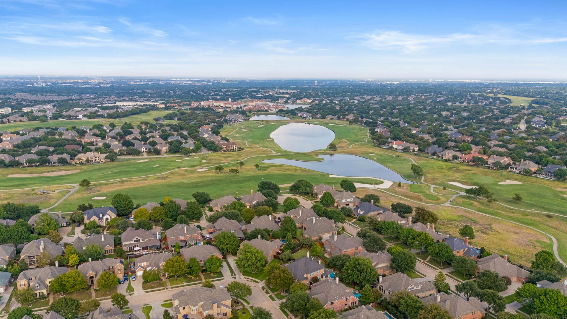 Aerial view of a suburban neighborhood next to a golf course with water features, houses with green lawns, trees, and a large city skyline in the distance under a blue sky with some clouds.