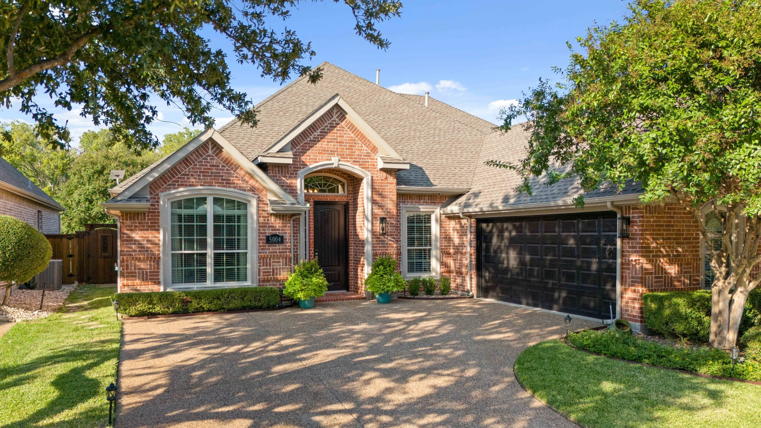 A brick house with a dark front door and a two-car garage, surrounded by a well-maintained yard with bushes, trees, and a concrete driveway.