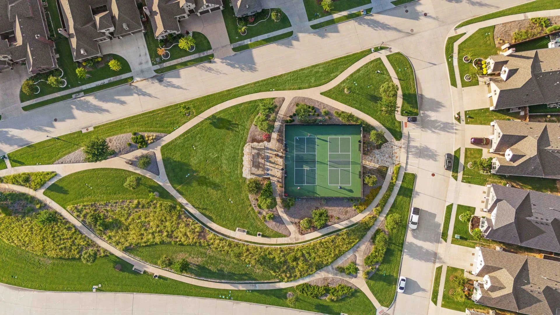 Aerial view of a residential neighborhood with houses, sidewalks, and a central green park area with trees, grass, walking paths, and a tennis court.