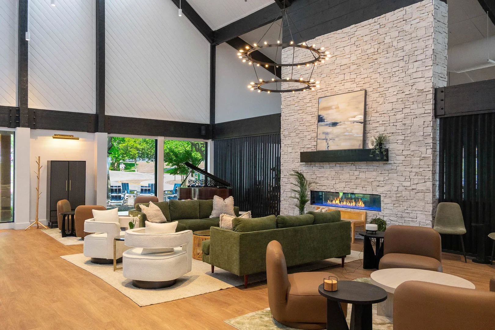 Interior of a modern living room with a high vaulted ceiling, large windows, and a stone fireplace, furnished with green sofas, chairs, and a piano visible near the windows.