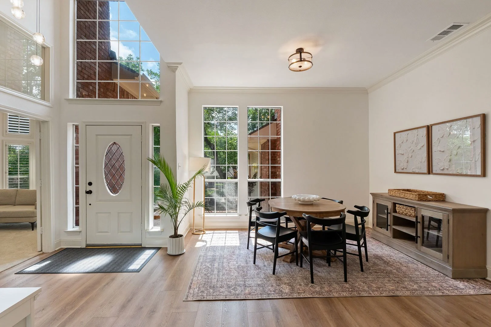 Bright and spacious living room with large windows, a round dining table with six chairs, a wooden sideboard, wall art, and a potted plant near the entrance.