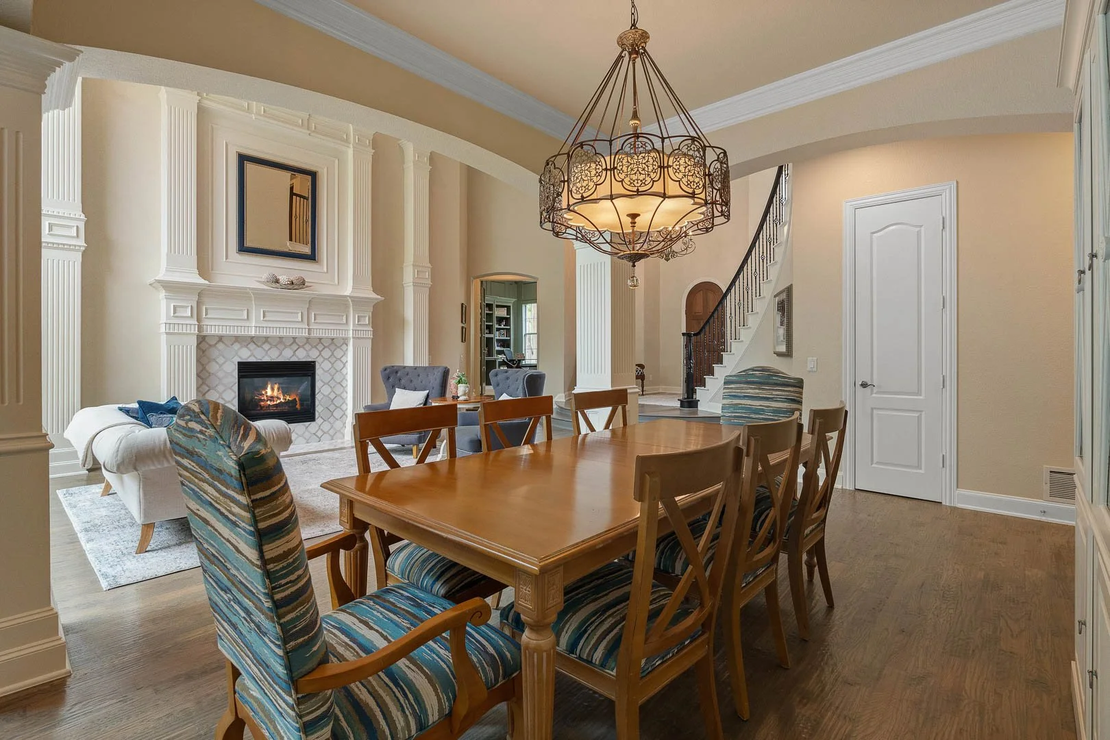 Dining room with a large wooden table, patterned chairs, a chandelier, and a living area with a fireplace and gray armchairs.