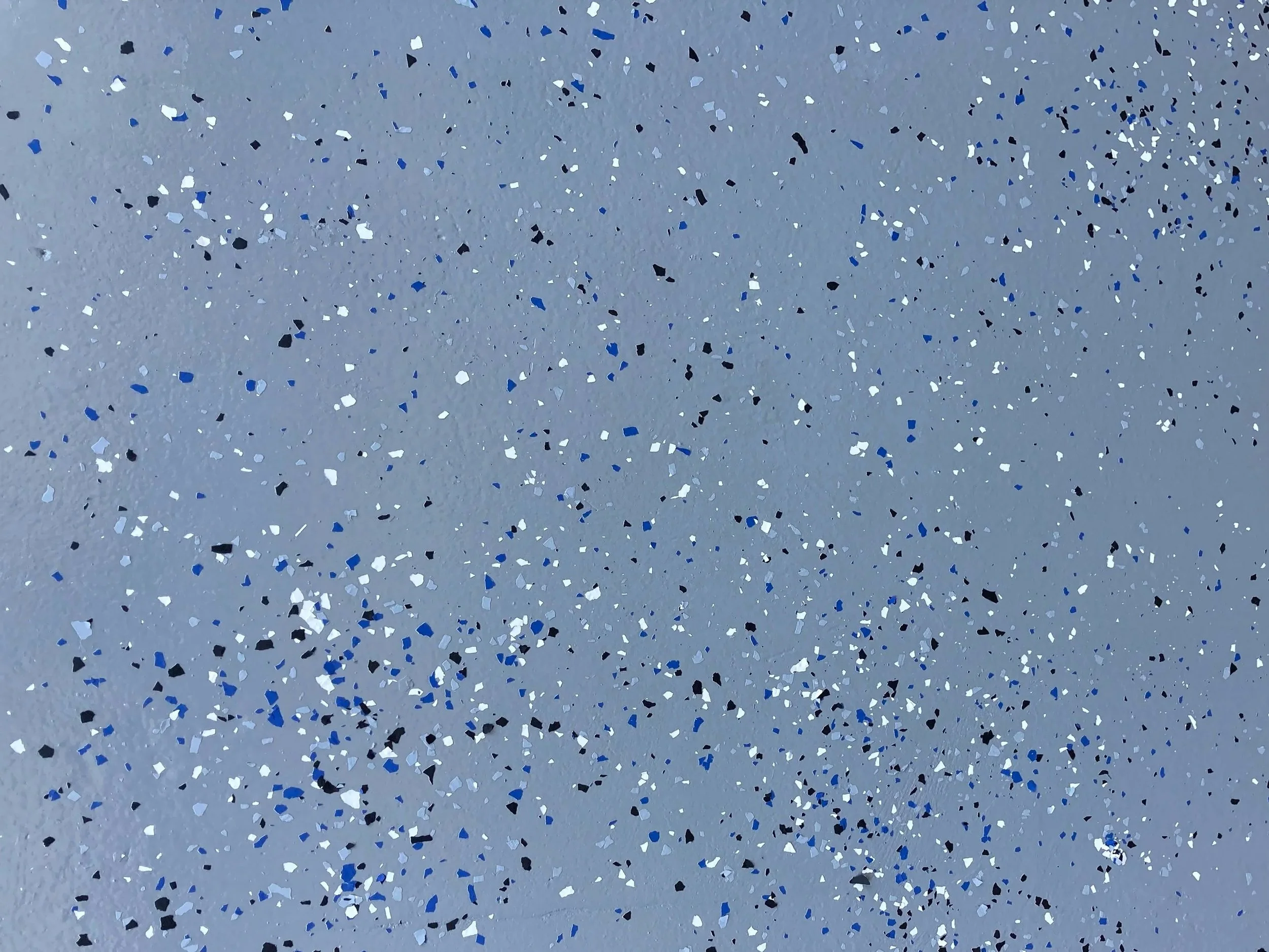Close-up of a gray epoxy floor, irregularly shaped blue, black, and silver glitter particles scattered across a light grey background.