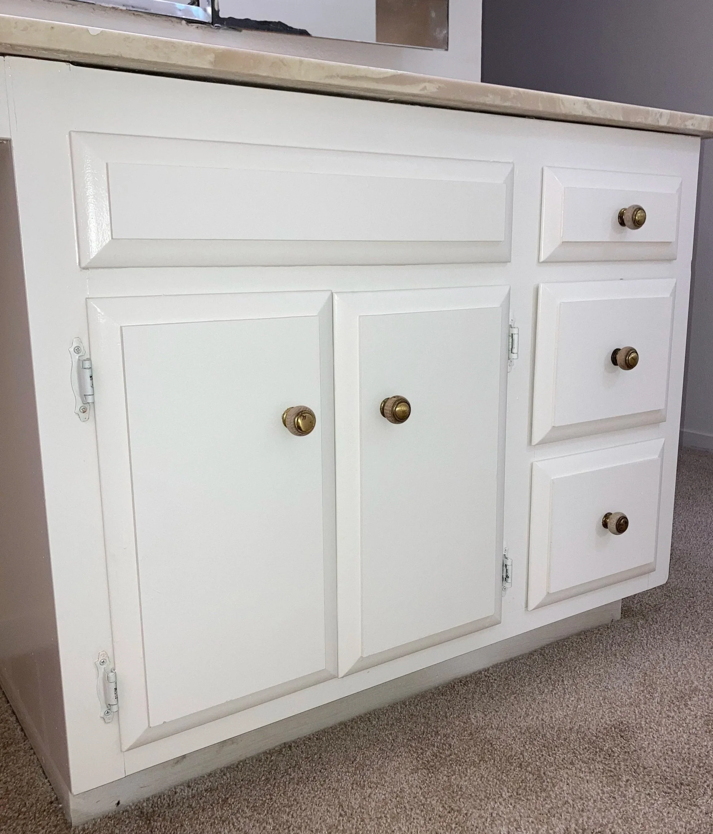 A freshly painted white bathroom cabinet with four paneled doors and four brass knobs, situated next to a beige carpeted floor and a beige countertop with a reflective surface.
