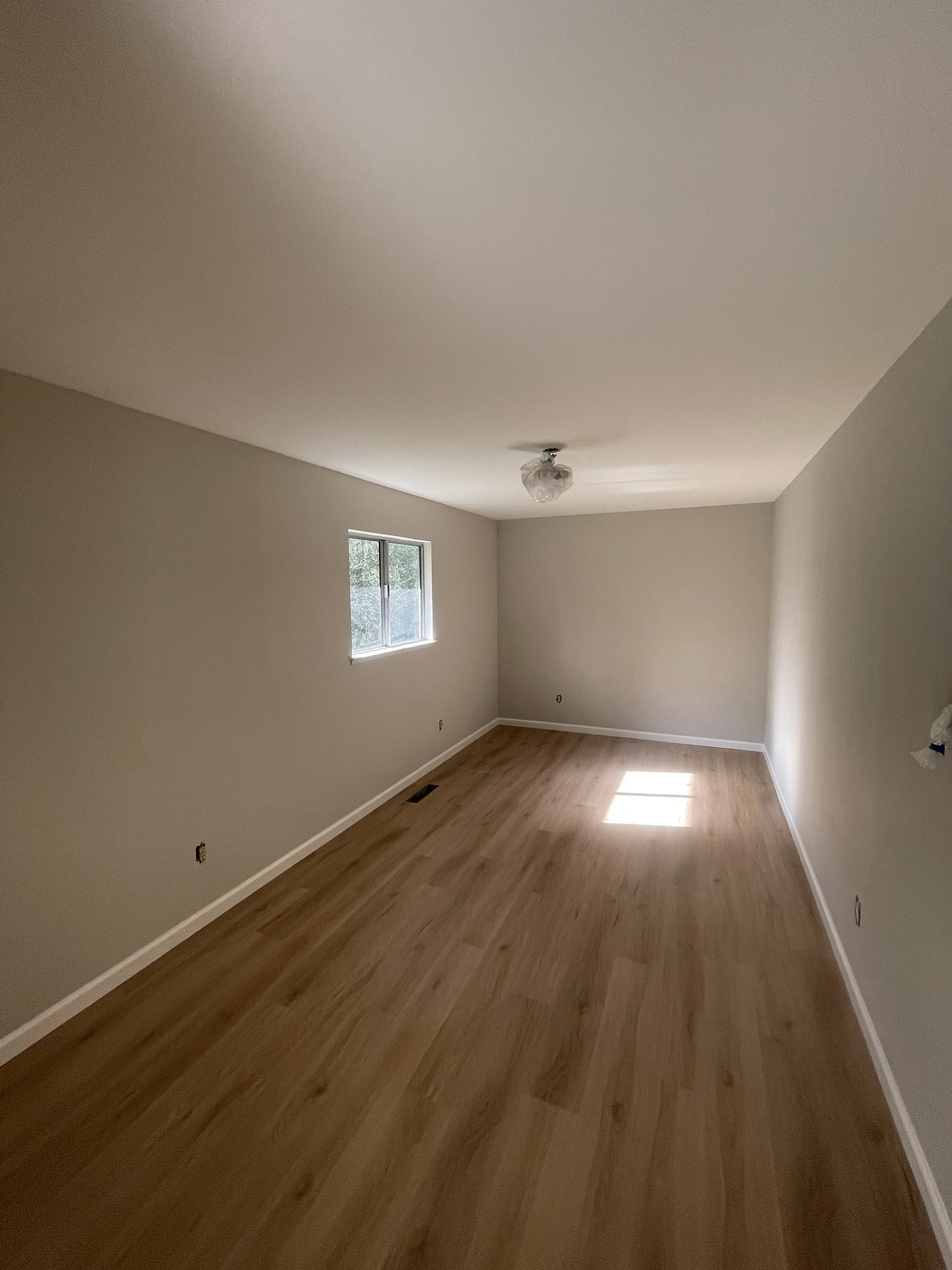 Empty room with newly painted, light-colored walls, hardwood flooring, a small window, and a ceiling light fixture covered with plastic.