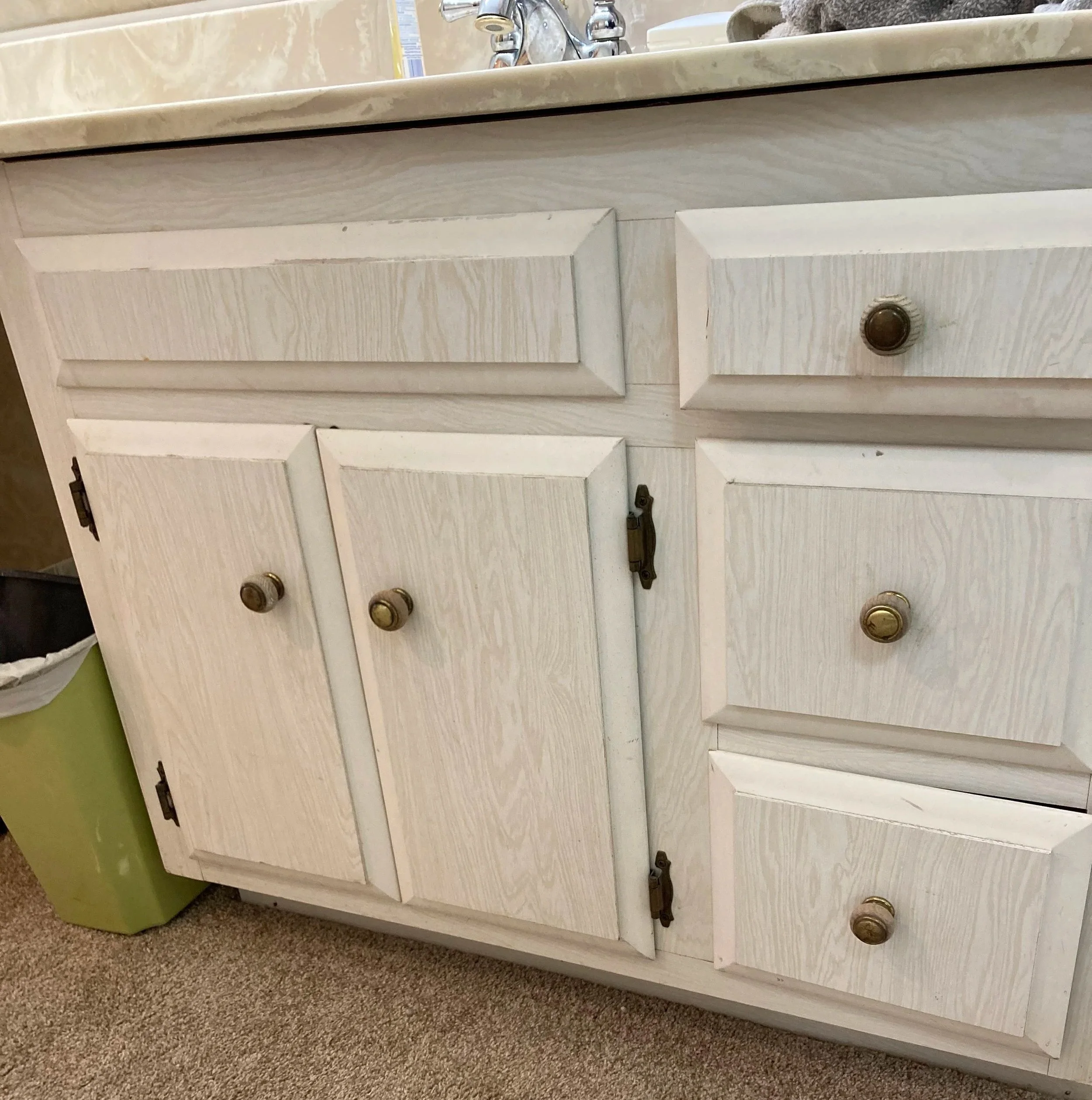 An outdated bathroom cabinet with three drawers and round brass knobs, situated on a brown carpeted floor, with a green plastic container partially visible to the left and a bathroom sink visible above.