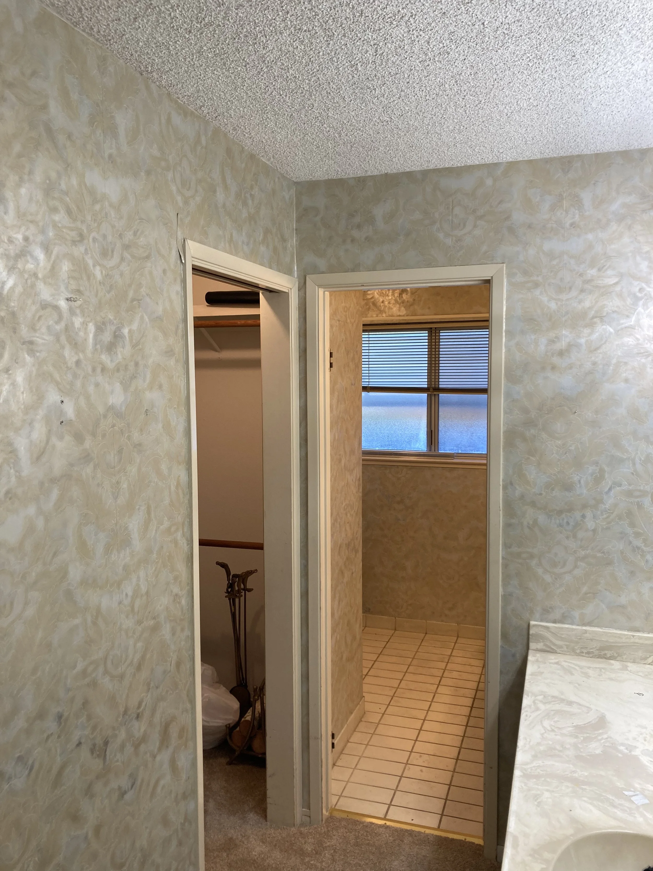 Interior view of a bedroom and bathroom with old, worn down textured wallpaper, a doorway leading to a sunlit room with a window and blinds, and a partially visible countertop or cabinet.