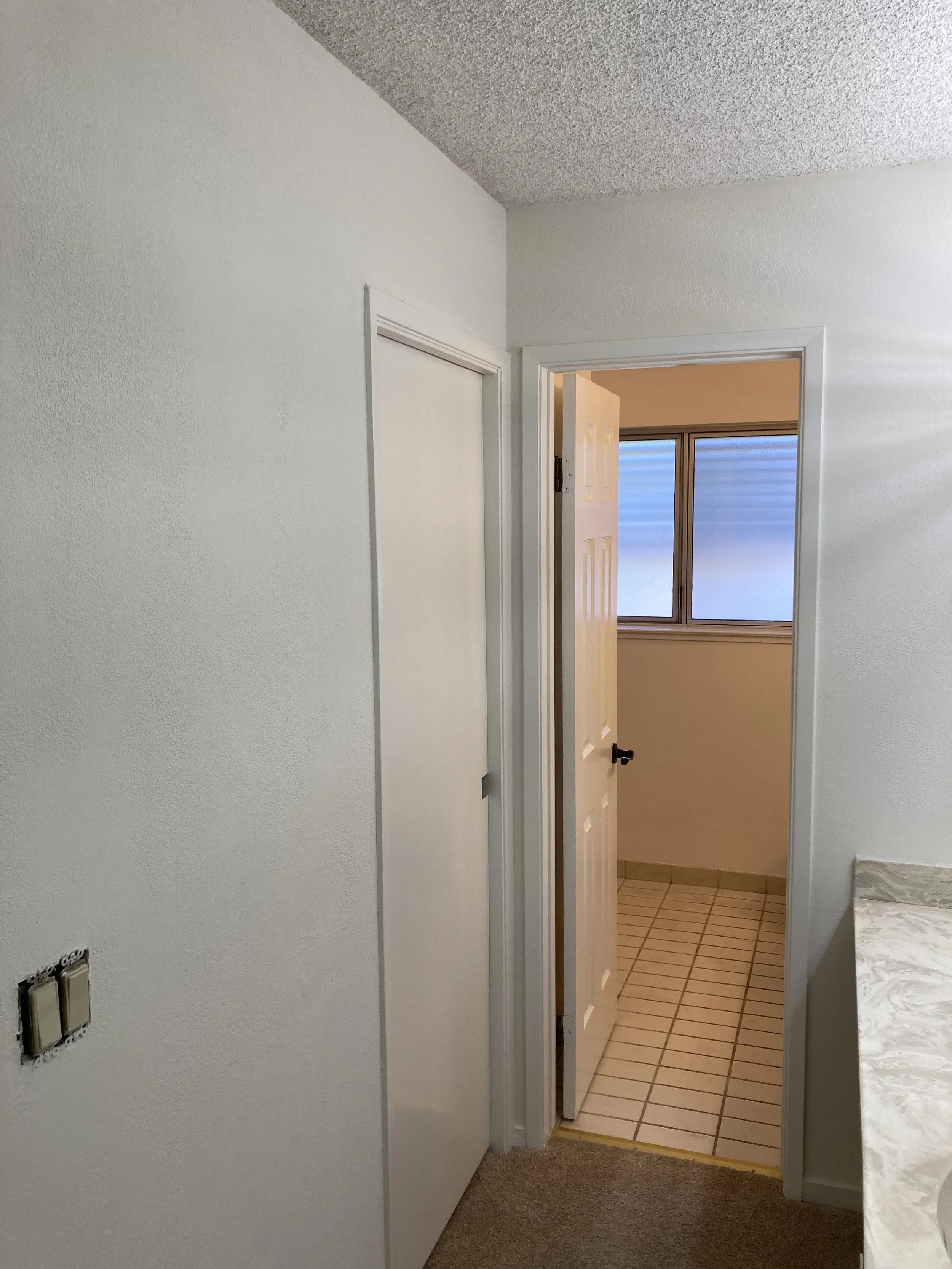 View of a newly painted hallway leading to a room with a window, beige walls, white door, and tiled floor. Part of a marble countertop is visible on the right.