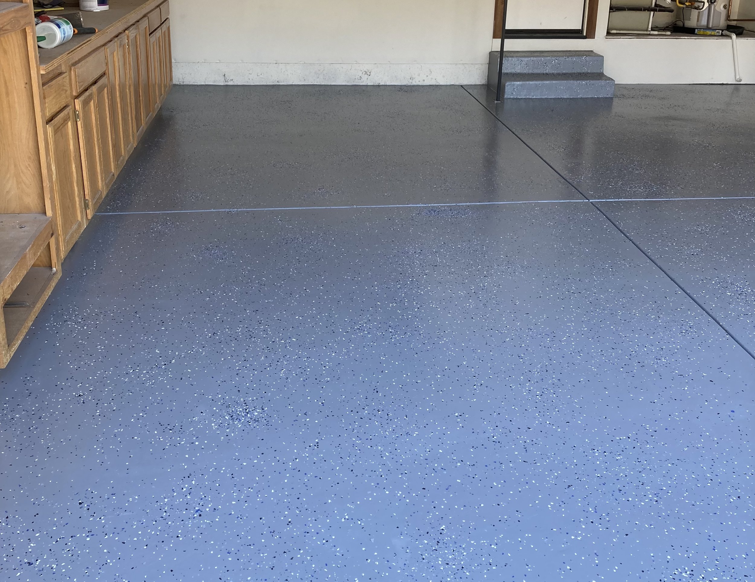 Garage with speckled gray epoxy floor, wooden cabinetry on the left, concrete stairs with a black railing, and a white wall with some cabinets and tools.