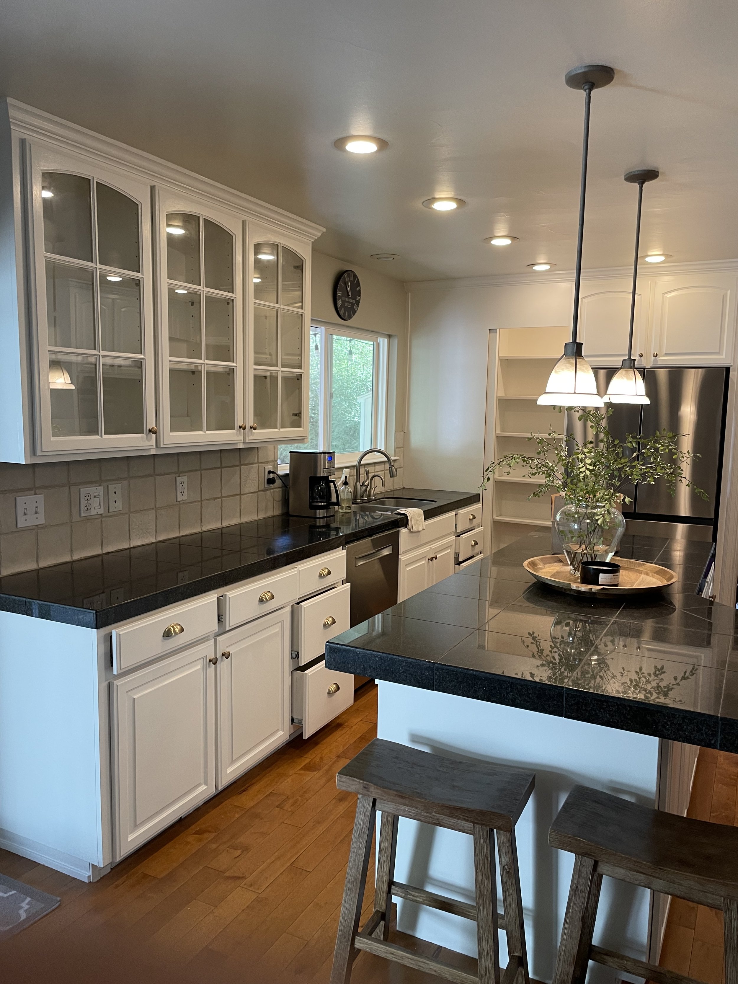 A kitchen with painted white cabinets, black countertops, a coffee maker, a window, a vase with greenery, pendant lights, and wooden stools.