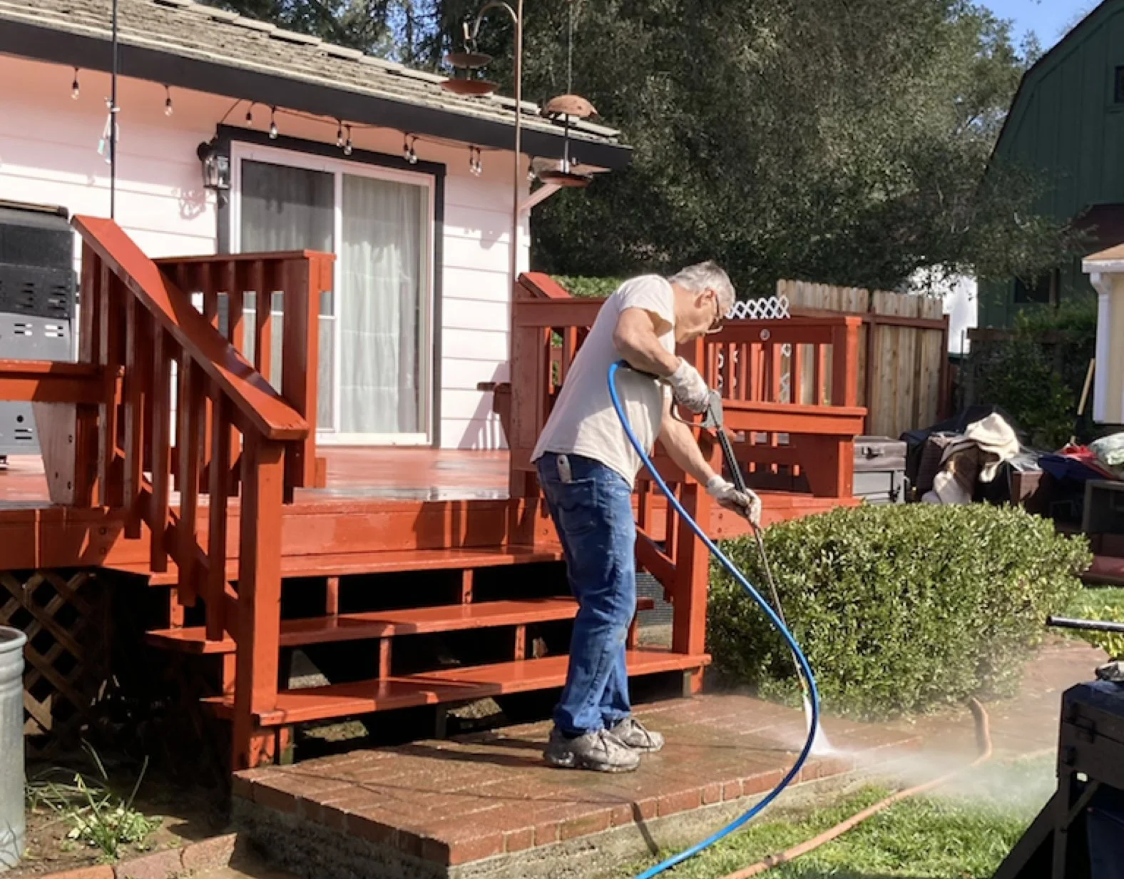 A man power-washing a wooden deck in a backyard, with a house in the background and a bush in the foreground.