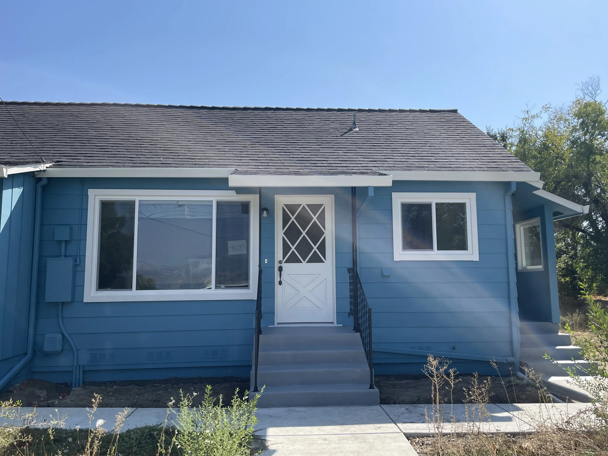 Front view of a light blue house with a dark gray shingled roof, white trim, and a white door with diamond-shaped windows, black handrails, and a small front porch.