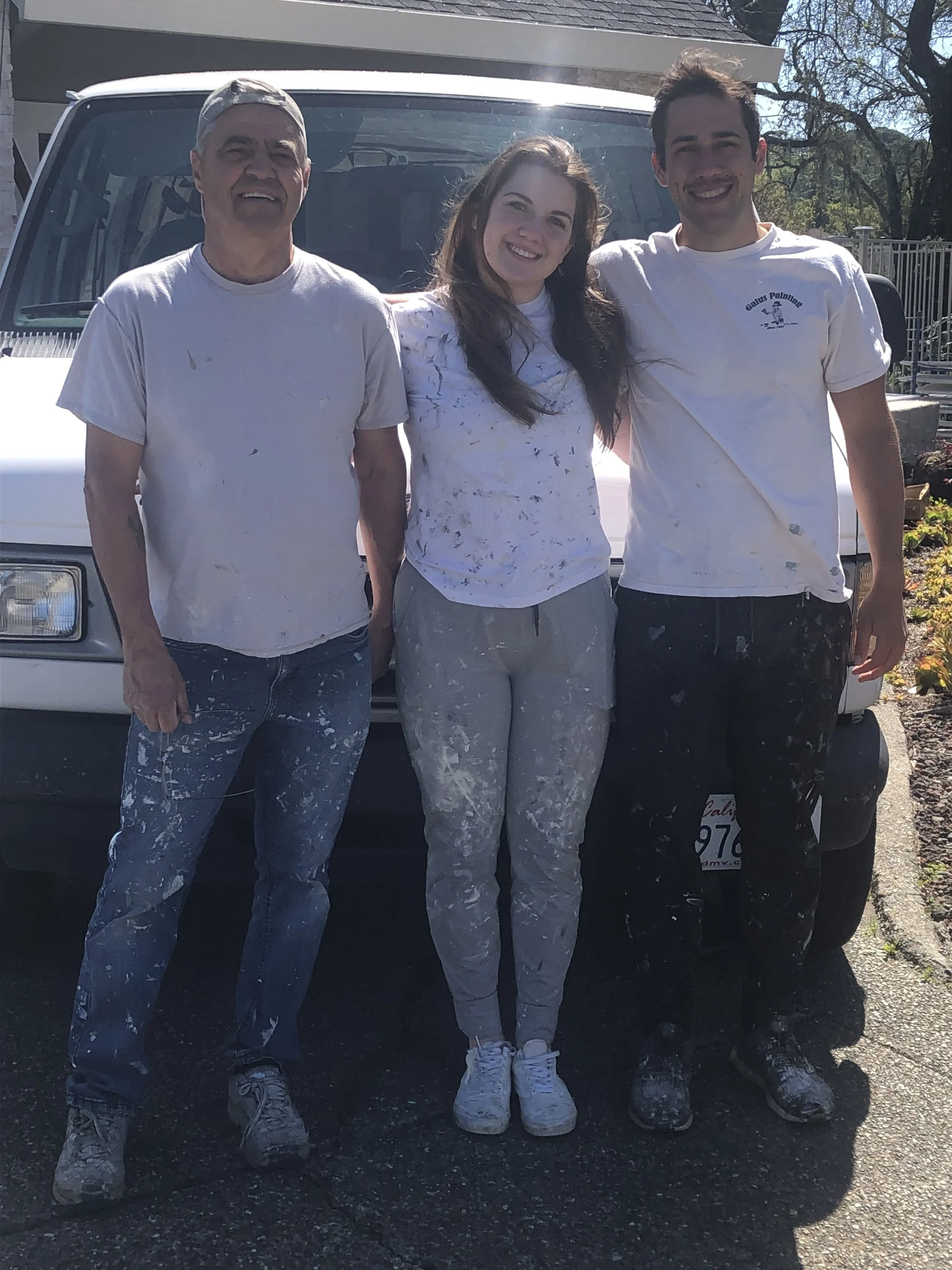 Three people standing in front of a vehicle, smiling, covered in white paint splatters, dressed casually in clothes suitable for painting.