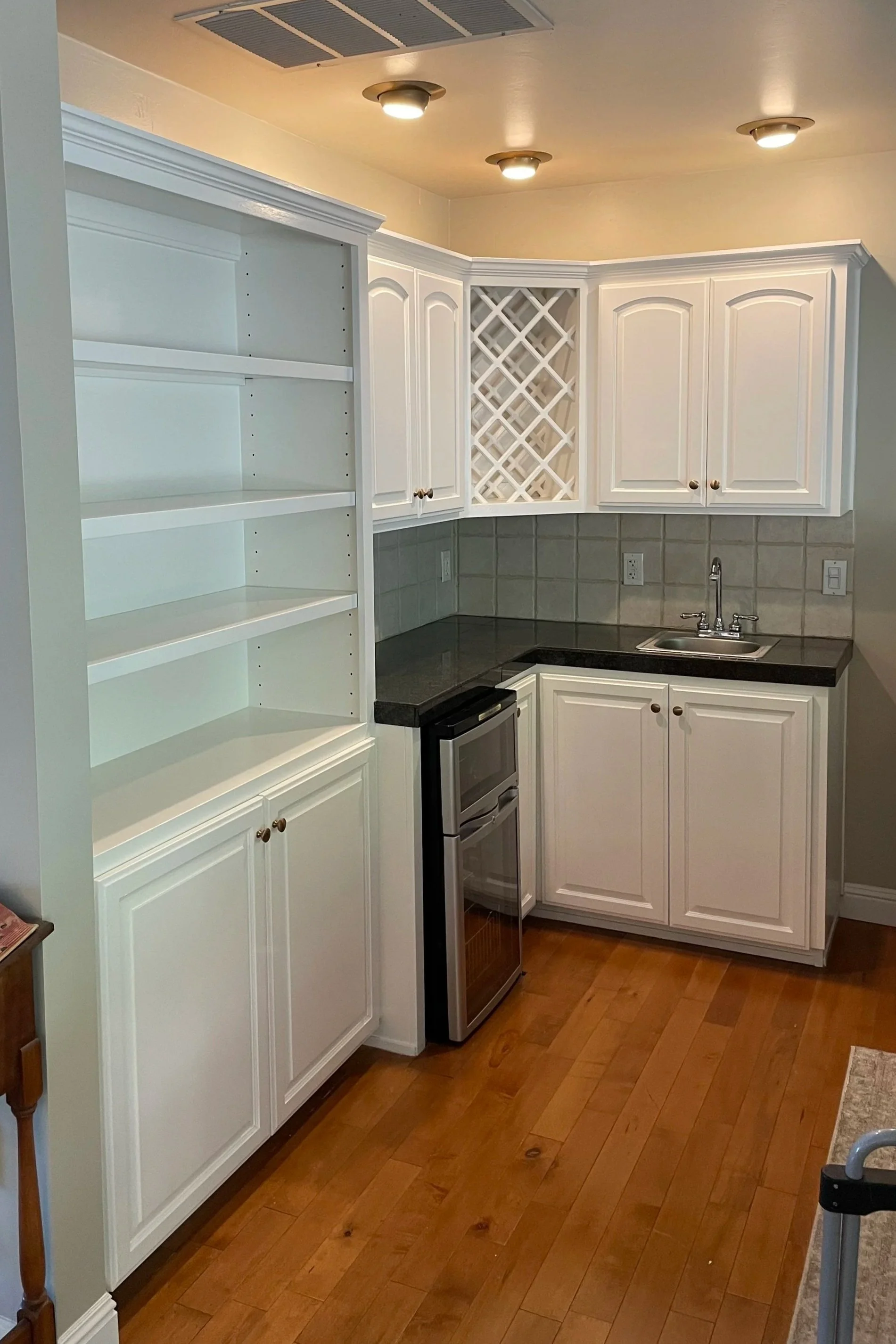 A recently painted kitchen with white cabinets, a black countertop, a small stainless steel fridge, a sink, and hardwood flooring.
